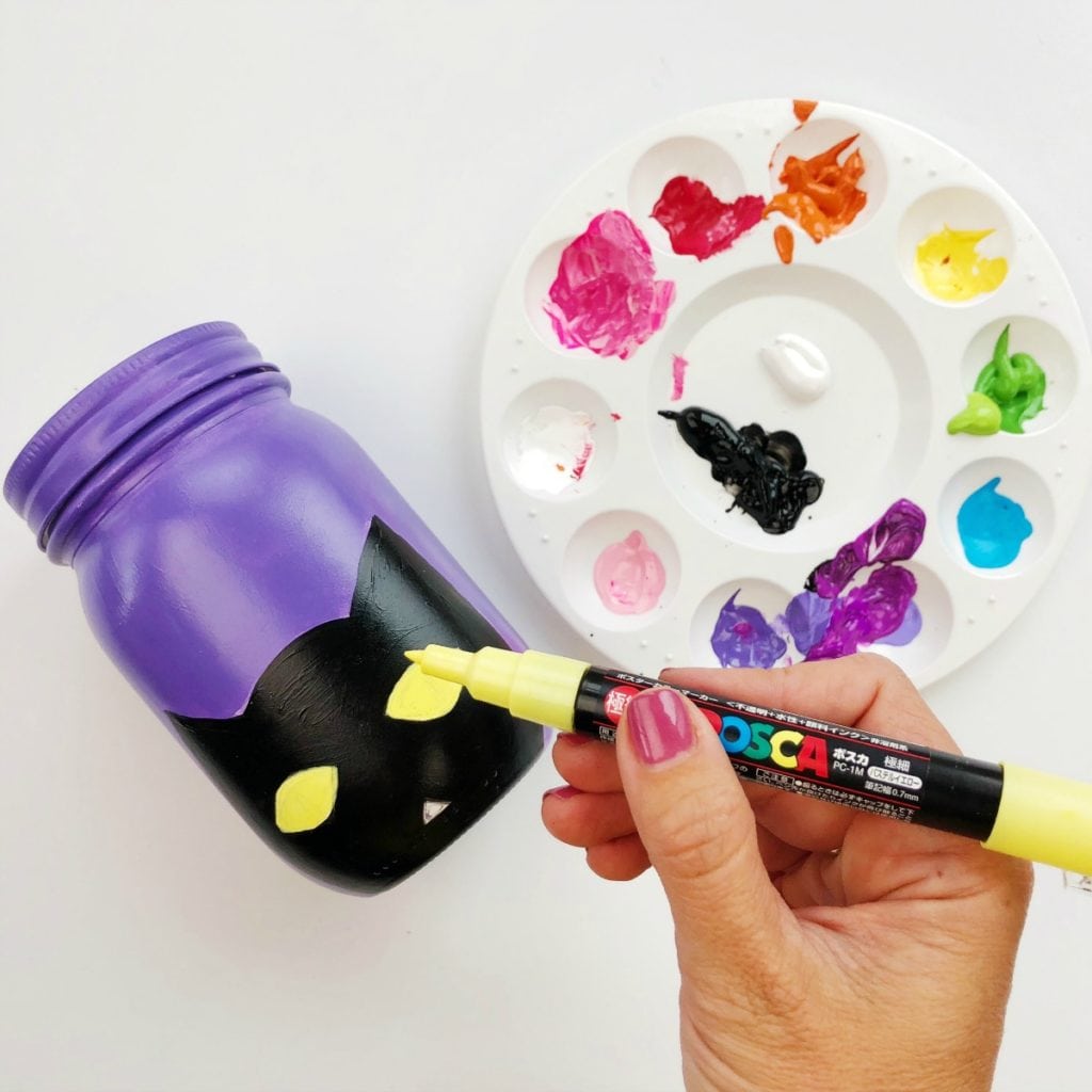Frasco roxo com um contorno de gato preto e os olhos sendo adicionados em amarelo com um marcador de tinta