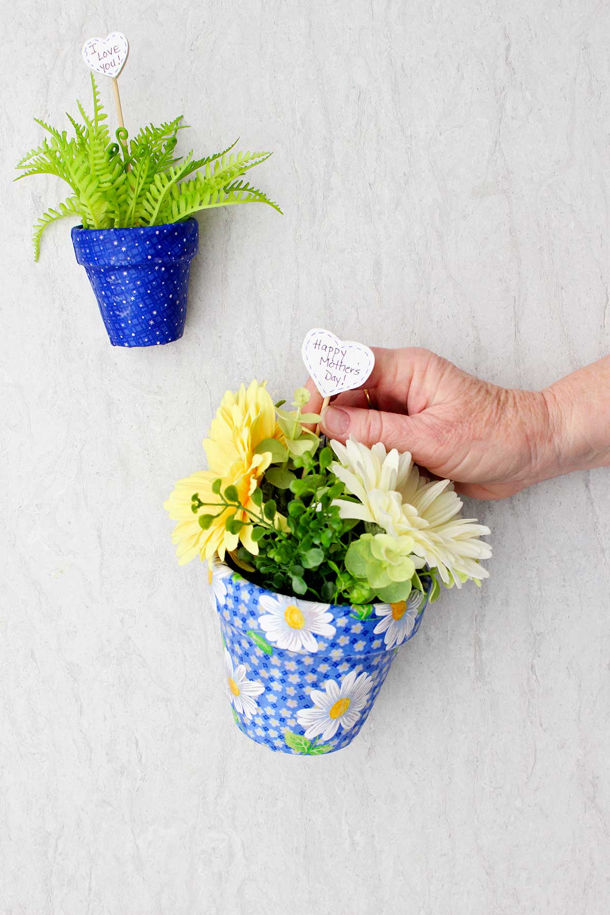 Uma pessoa colocando um post em forma de coração dizendo "Feliz Dia das Mães" dentro de um vaso de flores decoupado com flores.