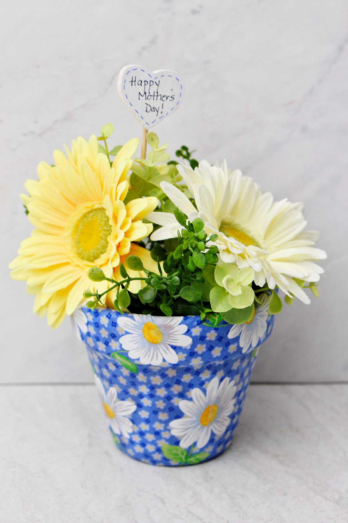 Um vaso decoupado com tecido azul de margaridas, com flores de primavera dentro e um post em forma de coração dizendo "Feliz Dia das Mães"