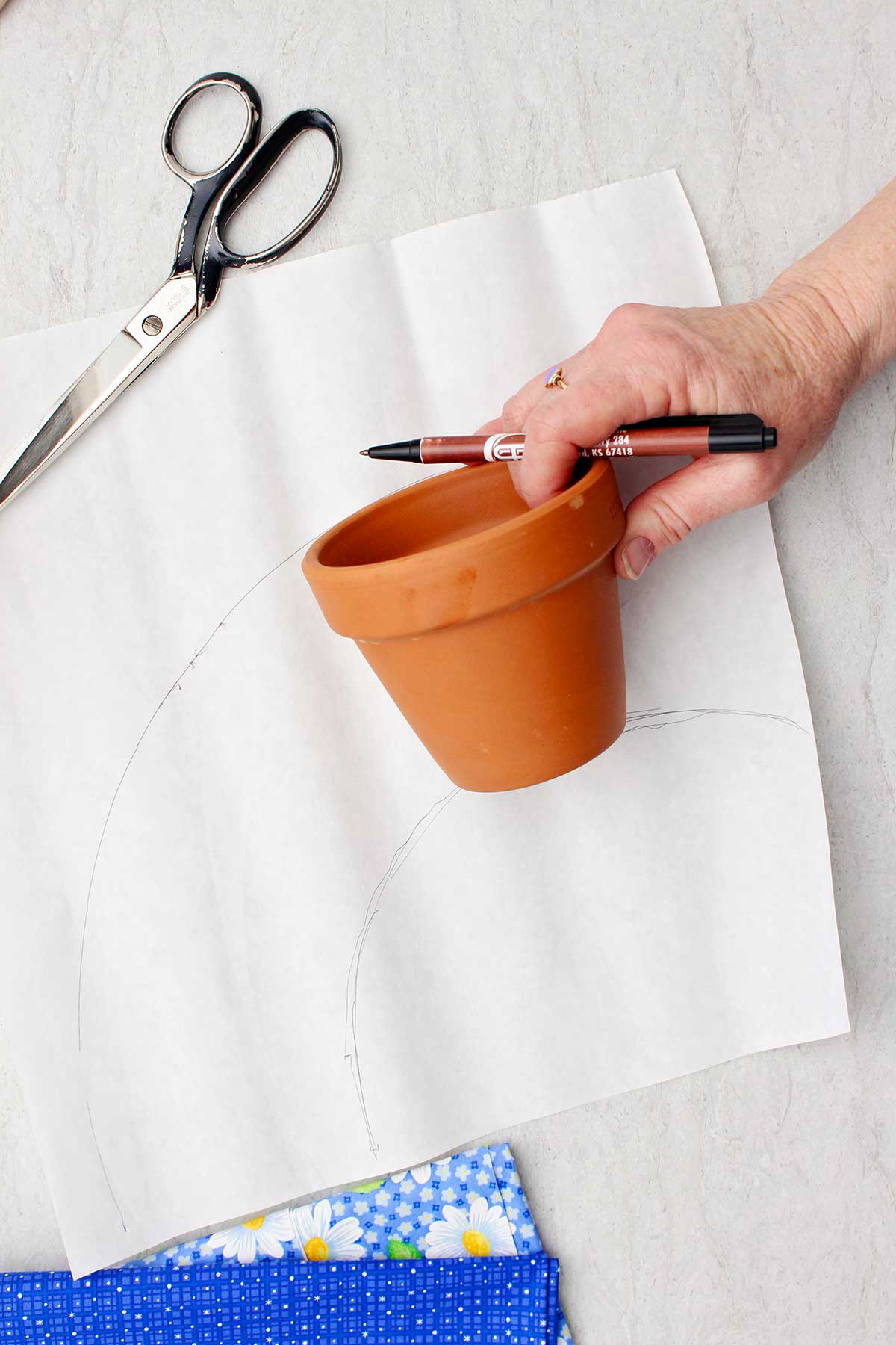 Uma pessoa traçando ao redor de um vaso de flores para ver quanto tecido ela precisará para cobri-lo.
