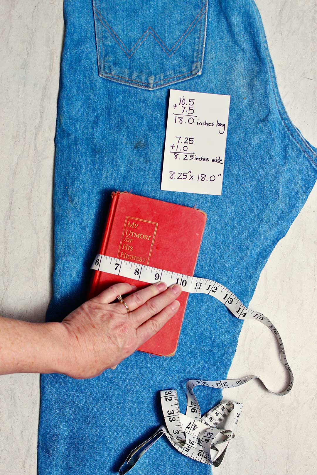 Uma mão medindo um livro para ver quanto tecido será necessário. Um cartão de índice mostrando medidas está sobre ele em um par de jeans velhos.