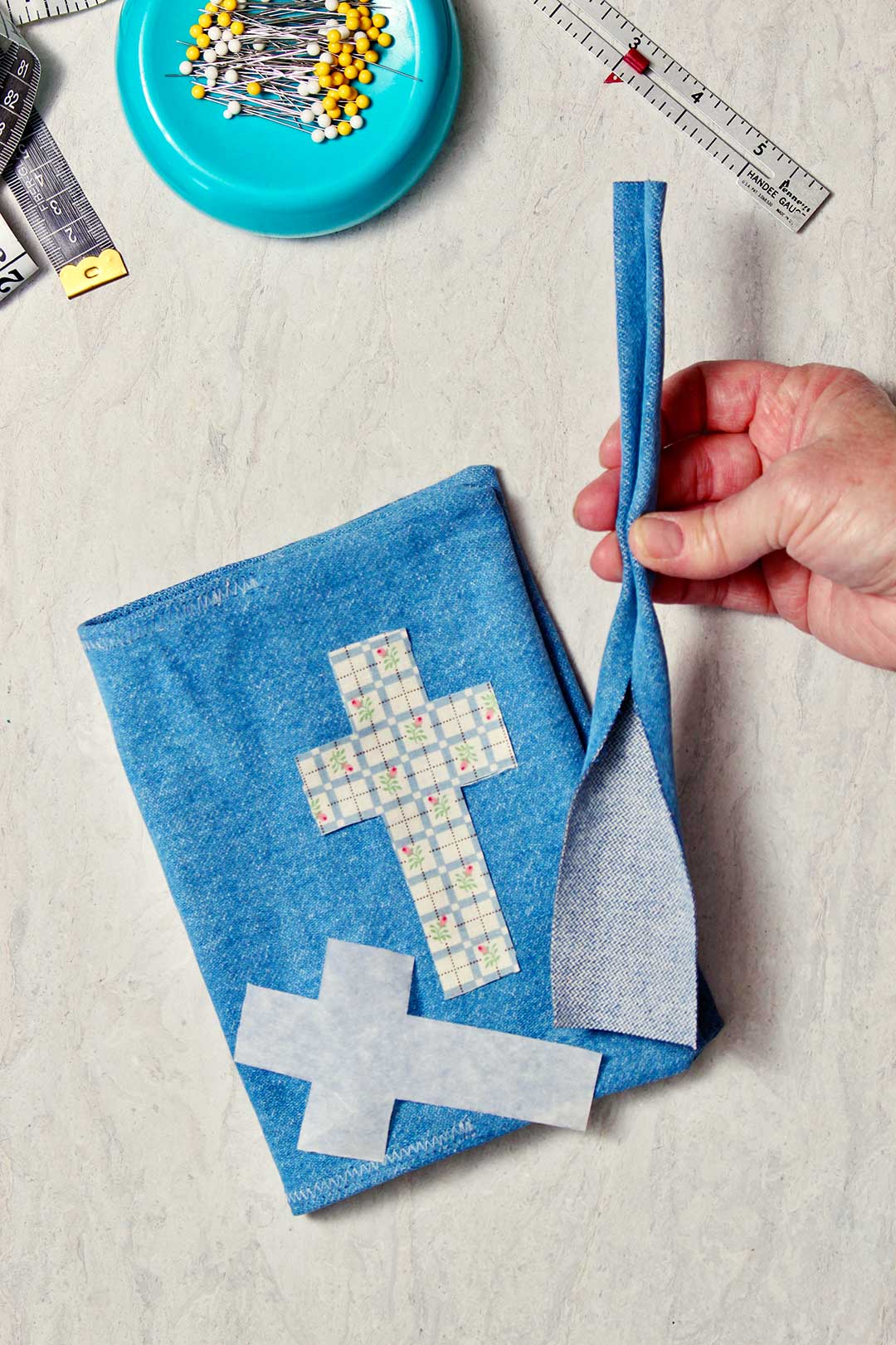 Uma mão segurando uma pequena tira de tecido de denim que será usada como alça para a capa de livro de denim.