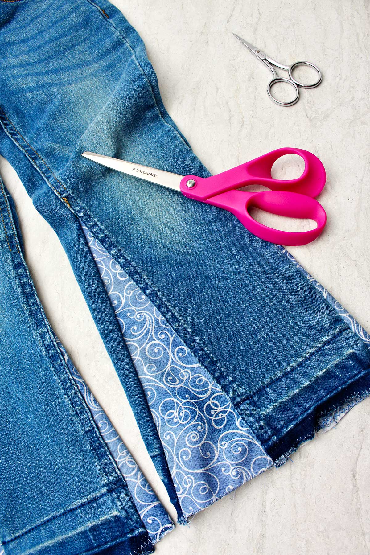 Um par de jeans do lado certo, com tecido decorativo costurado na lateral para fazer barra sino, com dois pares de tesouras por perto.
