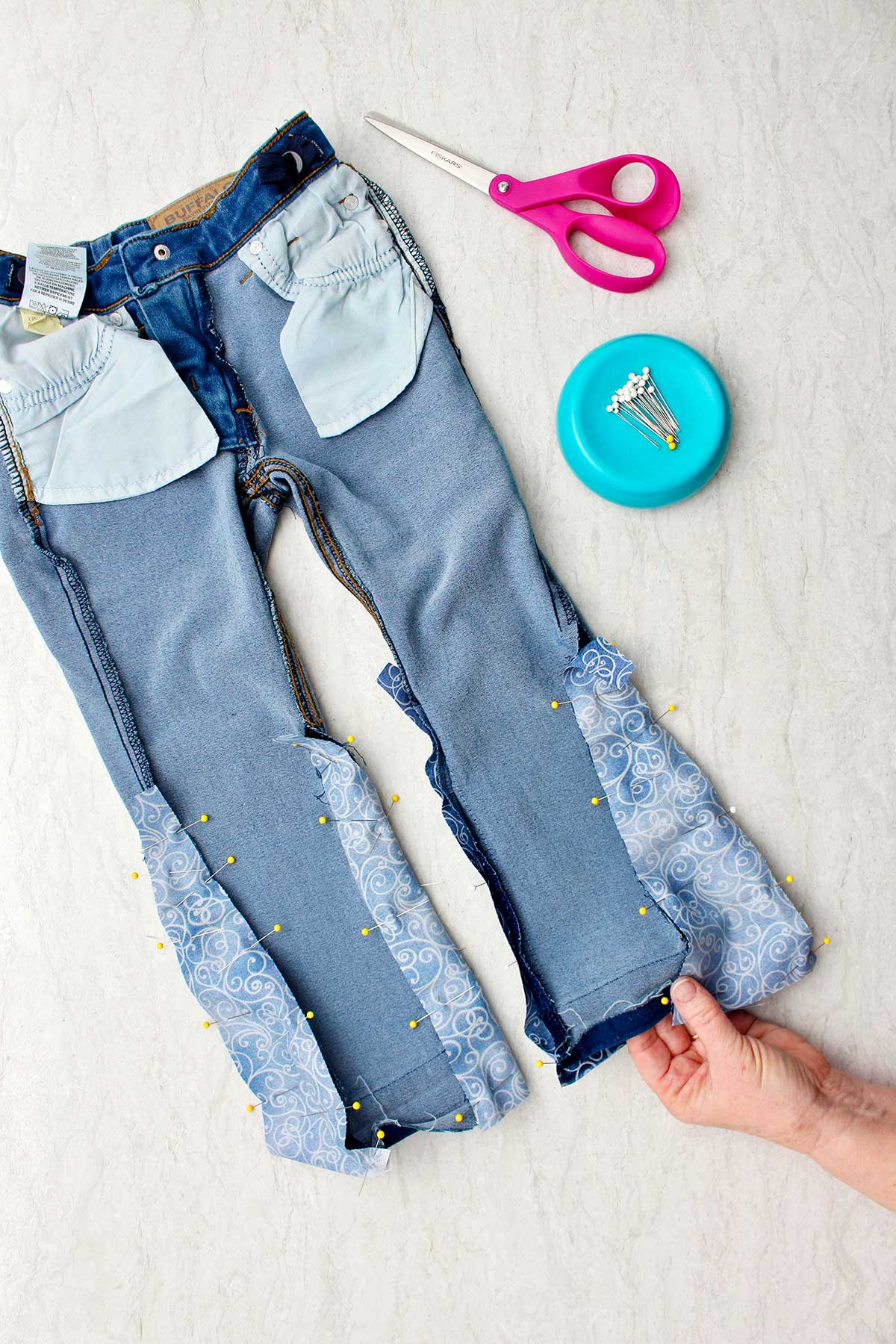 Jeans do avesso com alfinetes segurando o tecido no lugar para fazer barra sino, com alfinetes e tesouras por perto.