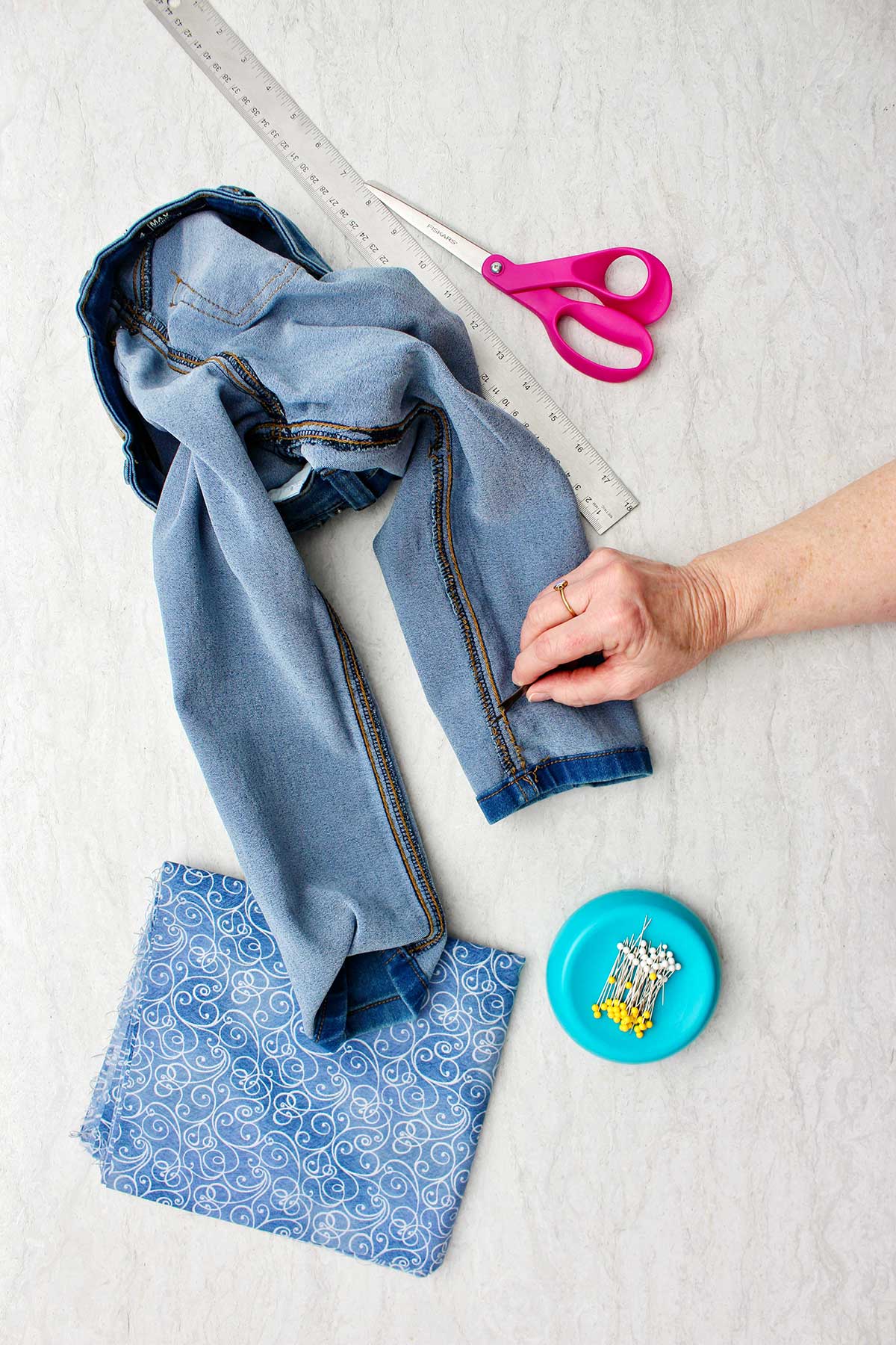 Um par de jeans do avesso, com a mão indicando onde ela quer cortar para fazer barra sino, com alfinetes, tesouras e tecido por perto.