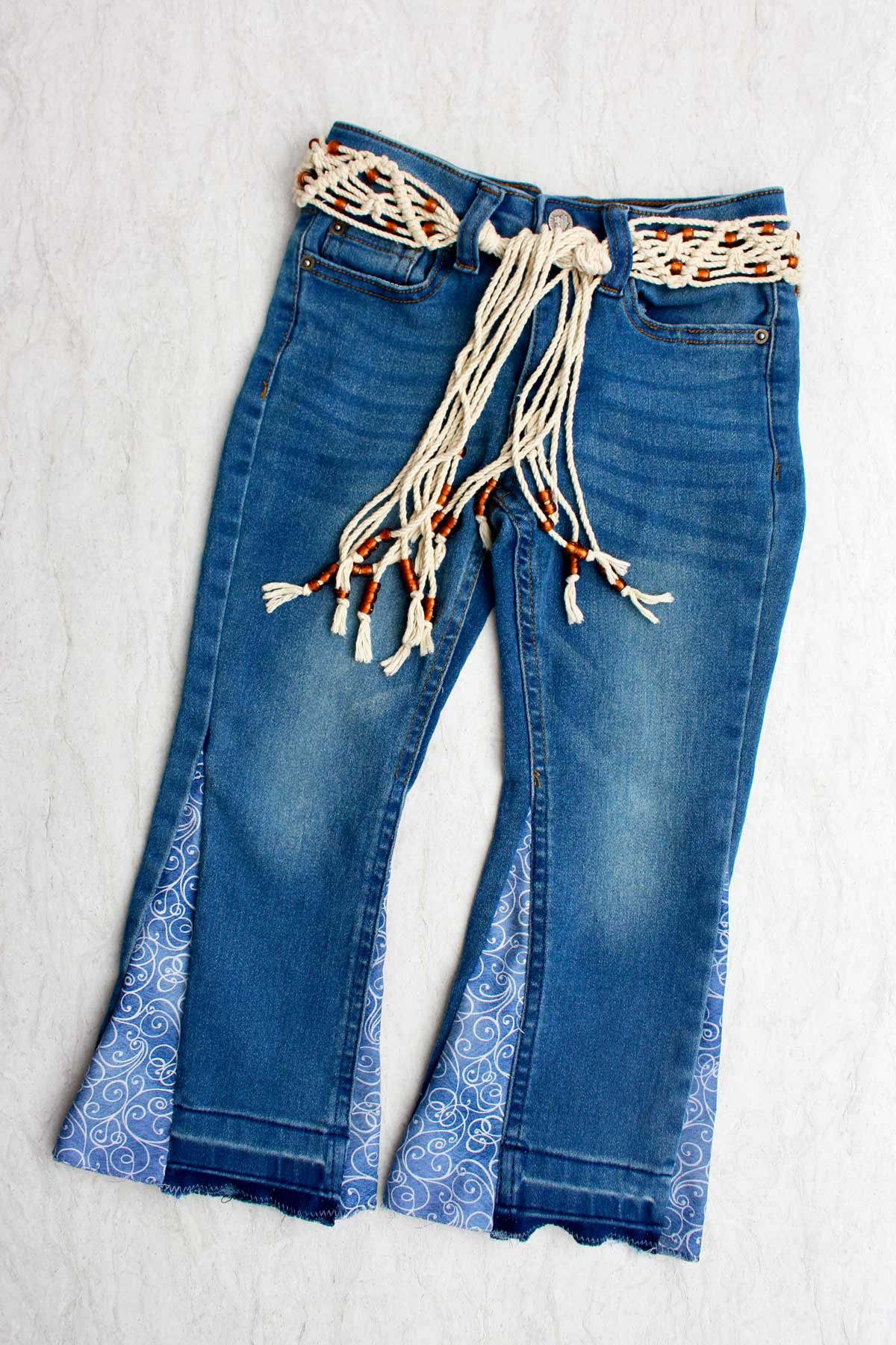 Um par de jeans infantis transformados em barra sino, com um tecido azul em espiral, presos com um cinto de macramê.