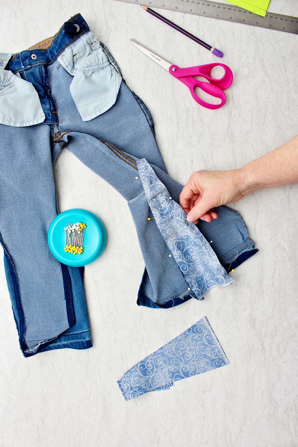 Mão segurando o tecido preso aos jeans com alfinetes retos, com suprimentos por perto.