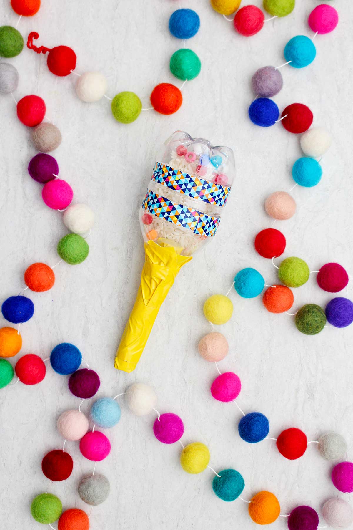 Maraca feita com uma pequena garrafa de água preenchida com arroz e contas, com um cabo amarelo e pompons coloridos por perto.