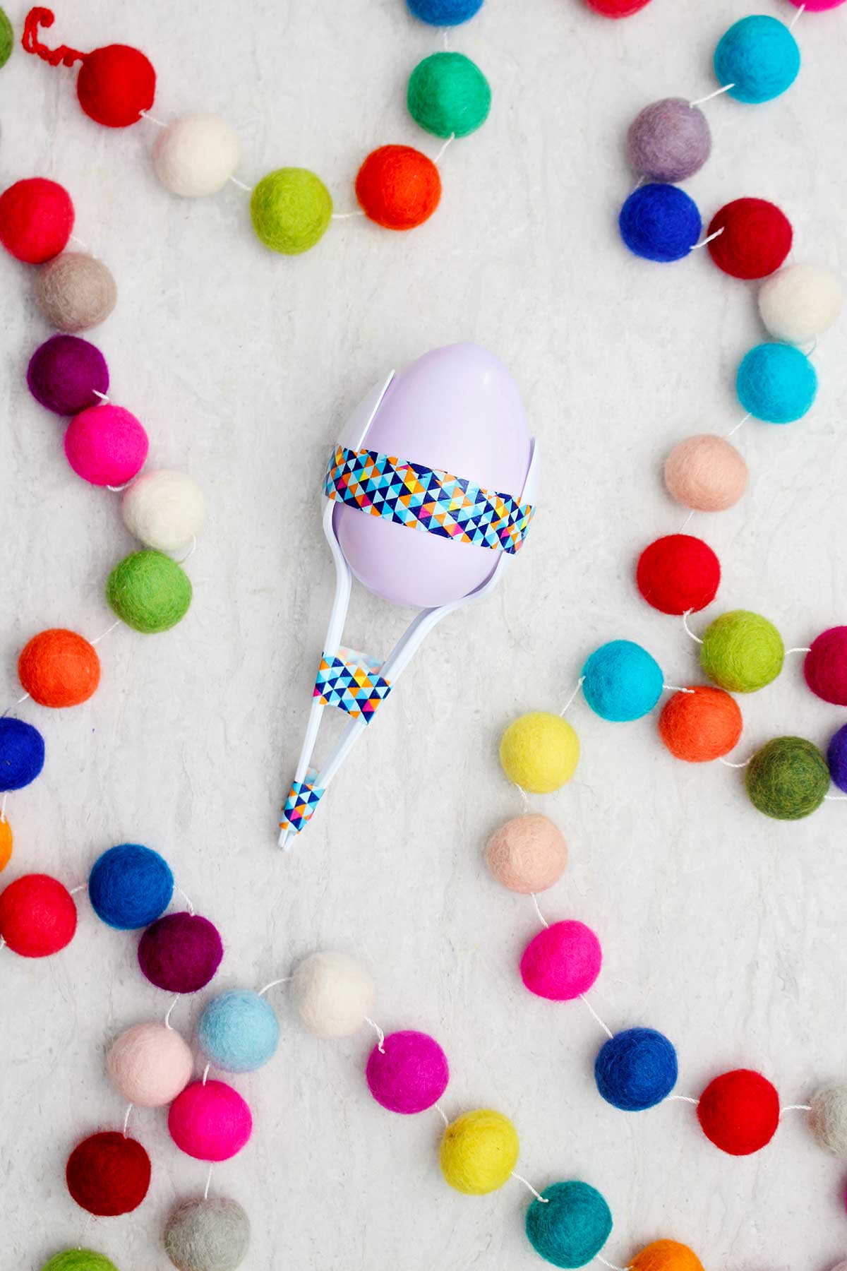Maraca de ovo de plástico lavanda com colheres e fita washi com pompons coloridos próximos.