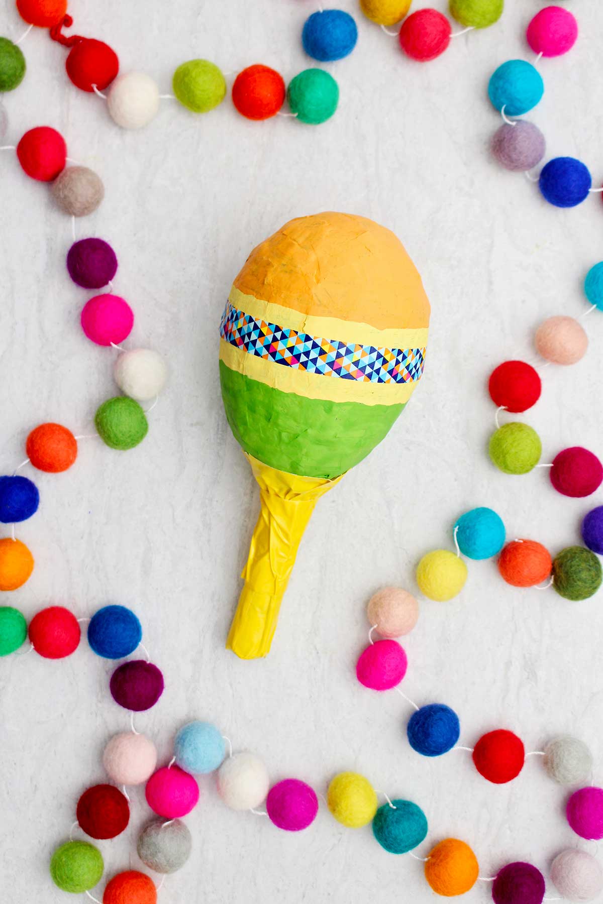 Maraca laranja, amarela e verde com cabo amarelo e fita washi colorida com pompons coloridos ao lado.