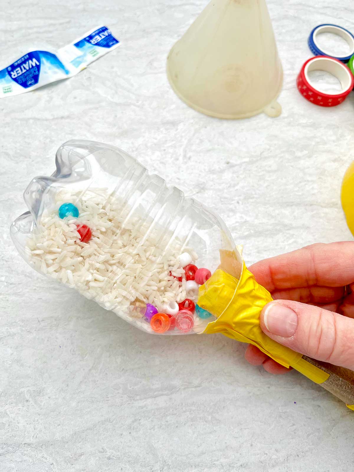 Pessoa segurando a maraca de garrafa com água para mostrar o arroz seco e as miçangas dentro.
