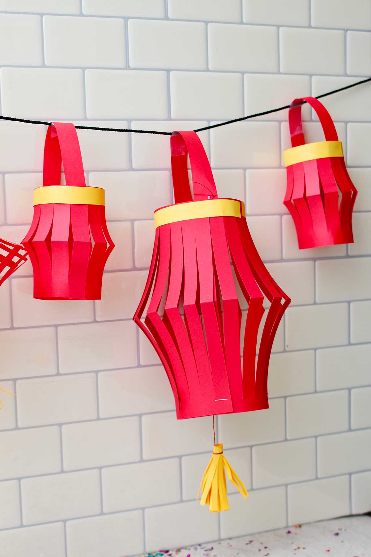 Três lanternas de papel completadas penduradas contra um fundo de azulejos de metrô.