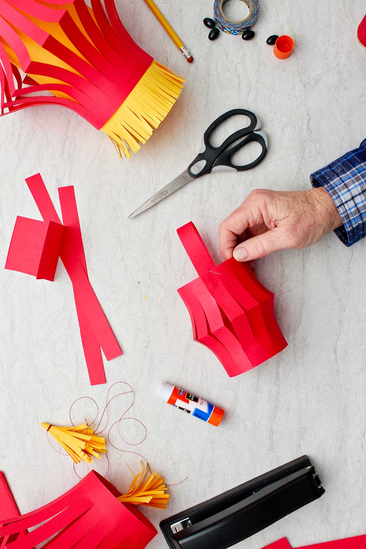 Mão segurando o corpo da lanterna de papel com uma tira de papel curvada sobre o topo com outros suprimentos ao redor.