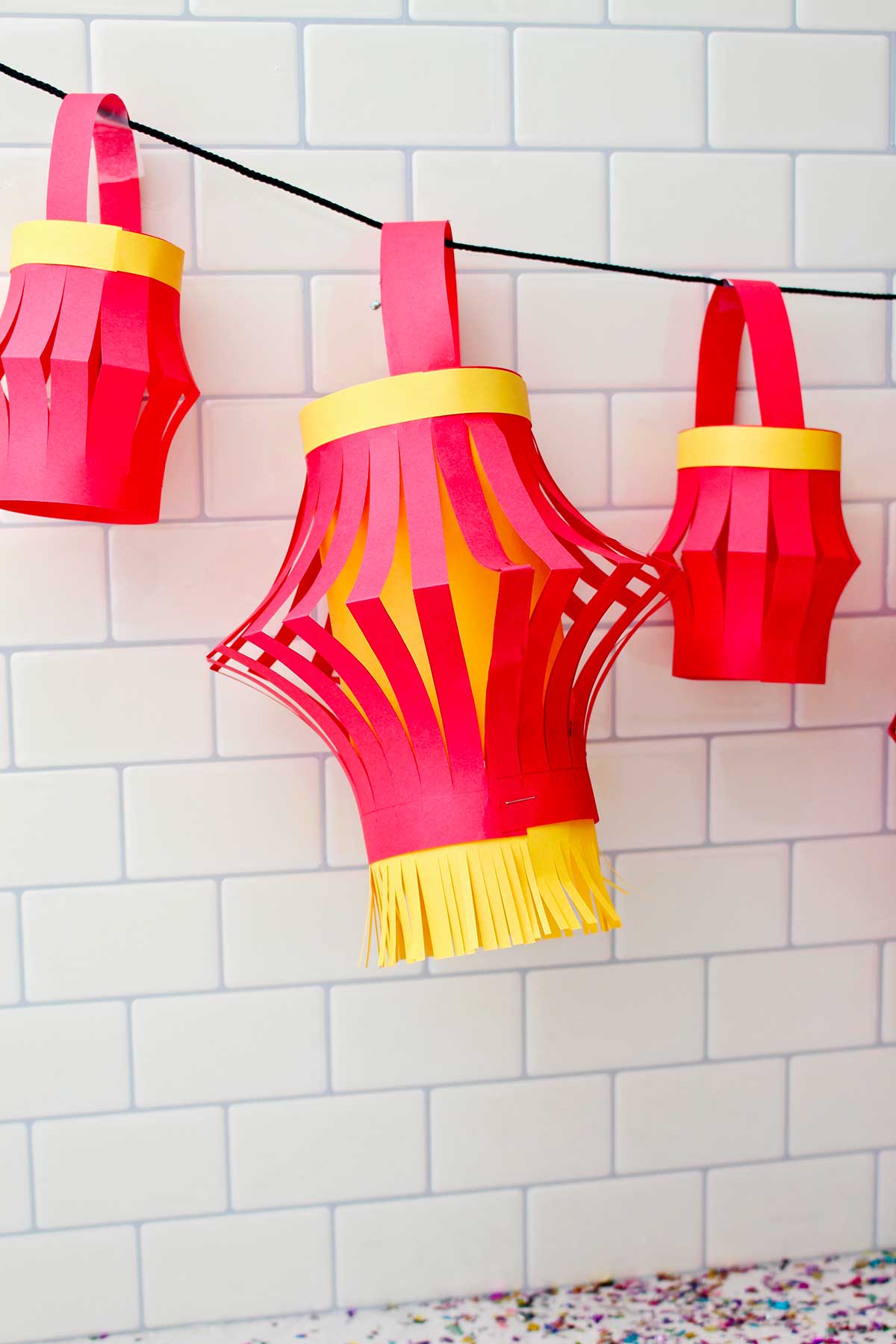 Três lanternas de papel completas penduradas contra um fundo de azulejos de metrô.