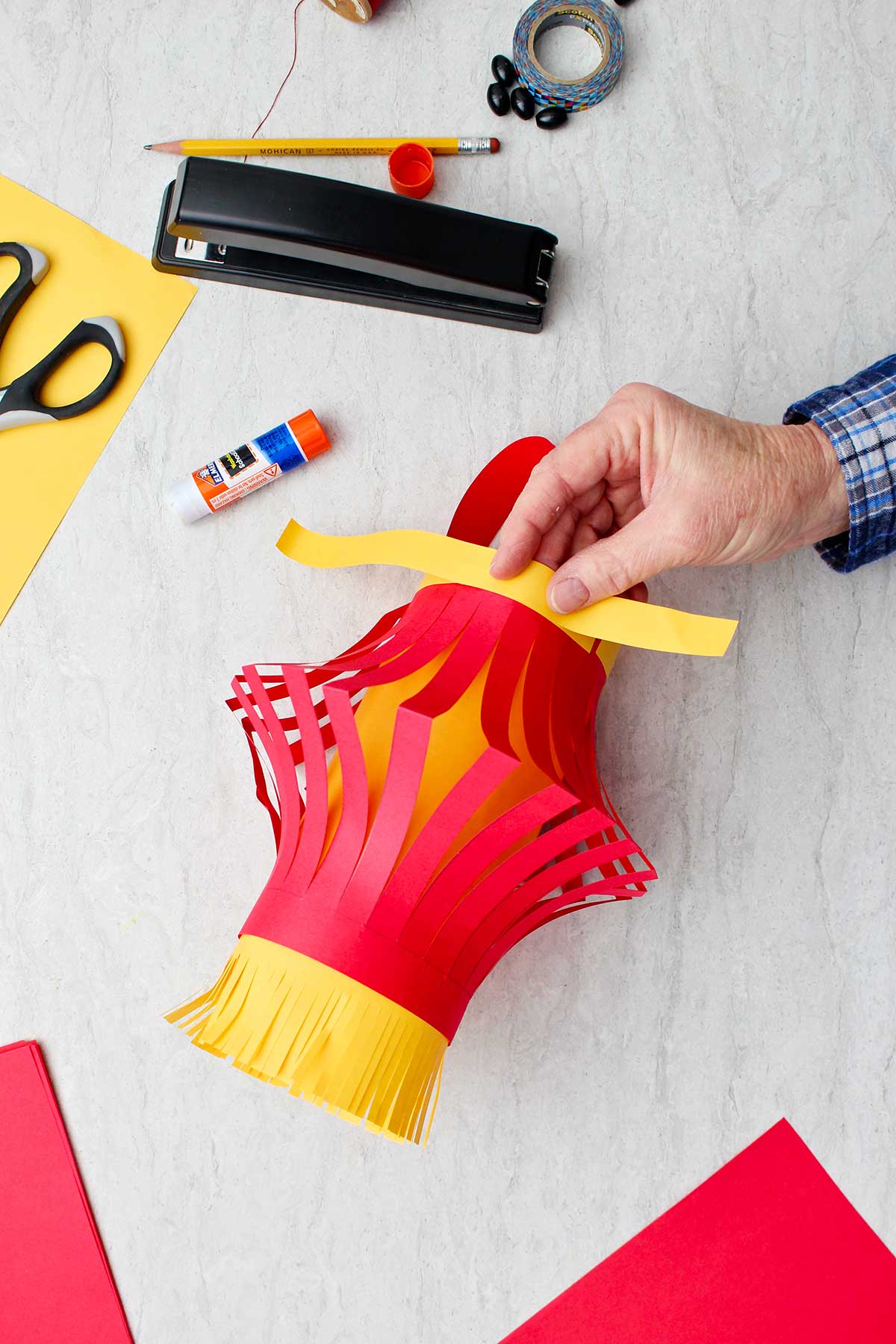 Mão colando uma tira de papel amarelo no topo da lanterna de papel para detalhes da decoração.