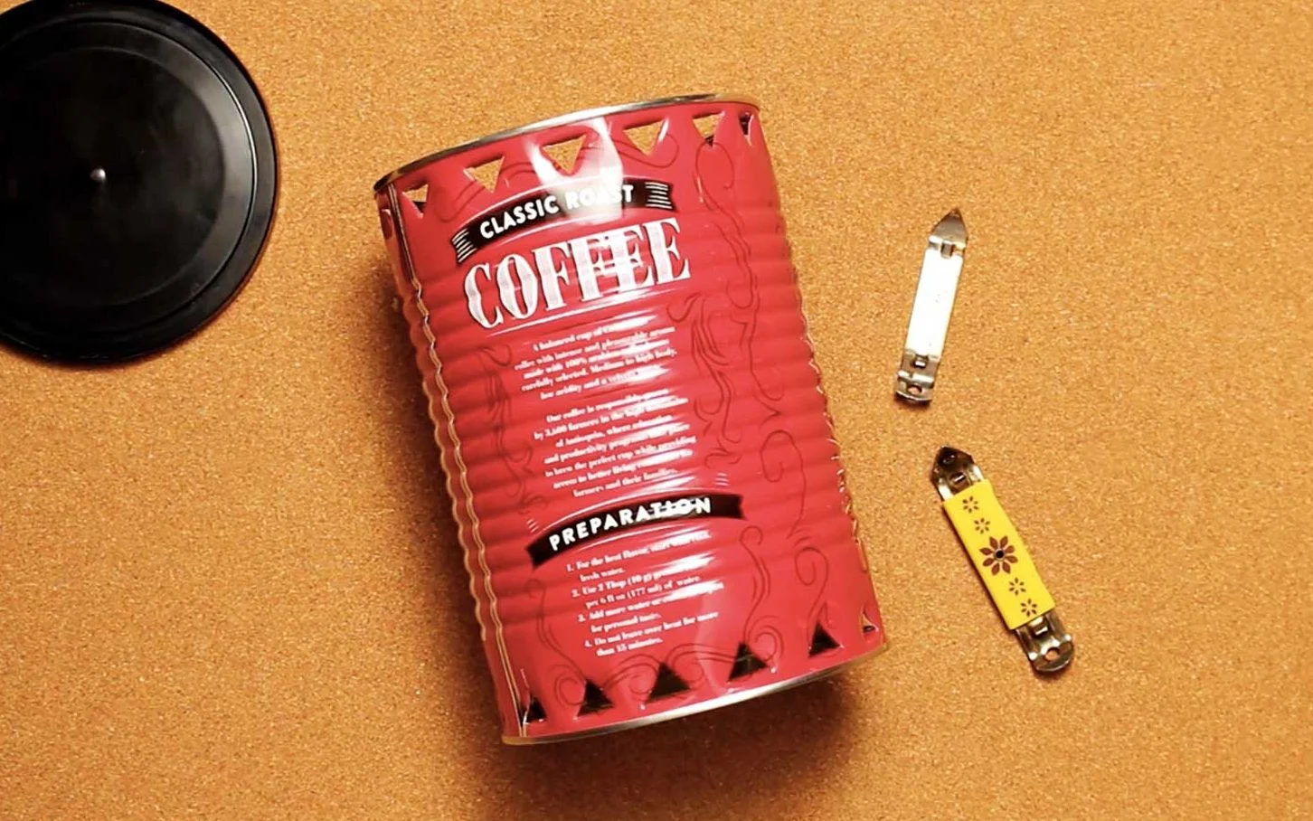 Uma lata de café, tampa e dois pequenos abridores de lata descansando sobre um fundo cor de cortiça.