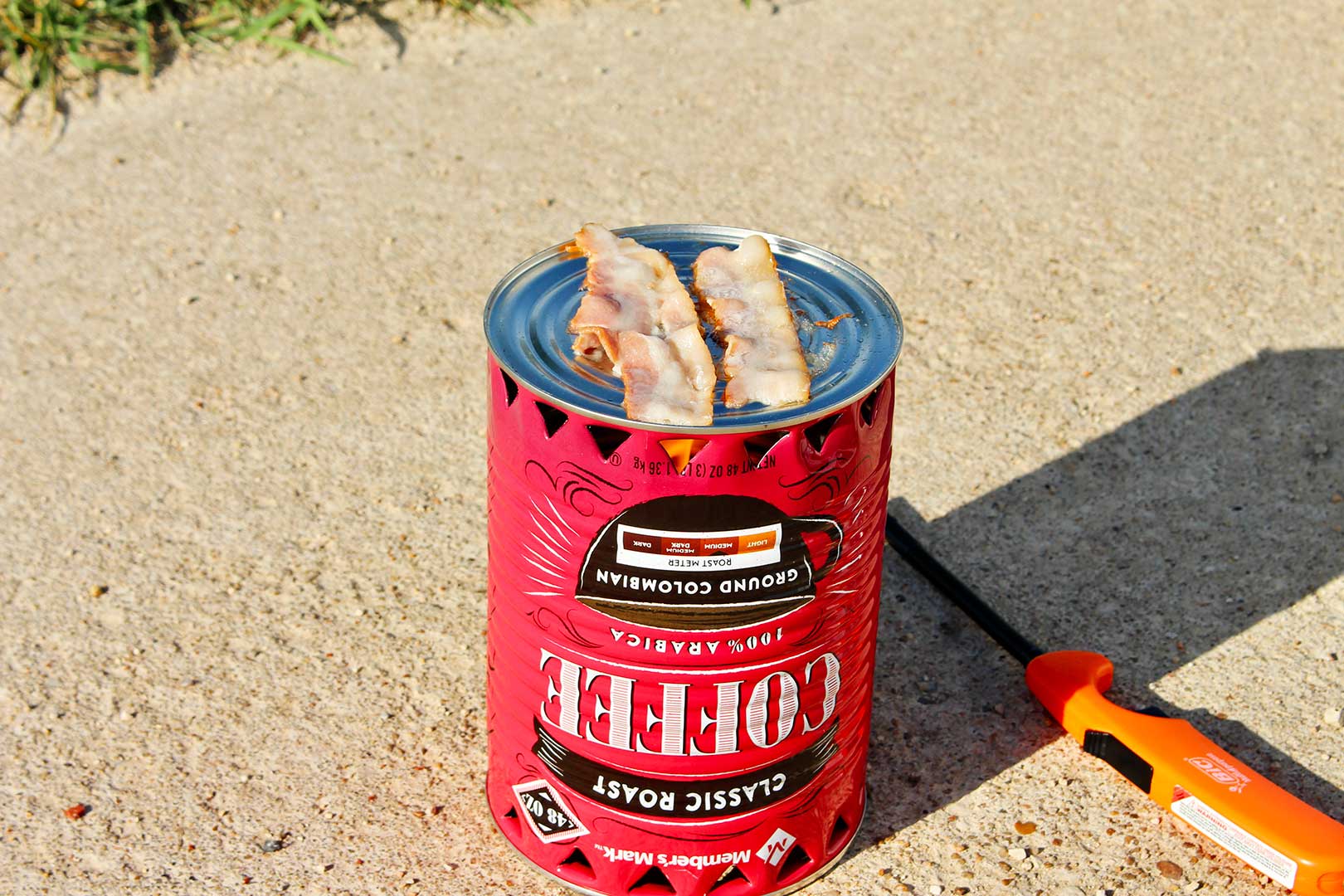 Pedaços de bacon prestes a serem cozidos em um fogareiro de pobre com um isqueiro laranja descansando na entrada da garagem.