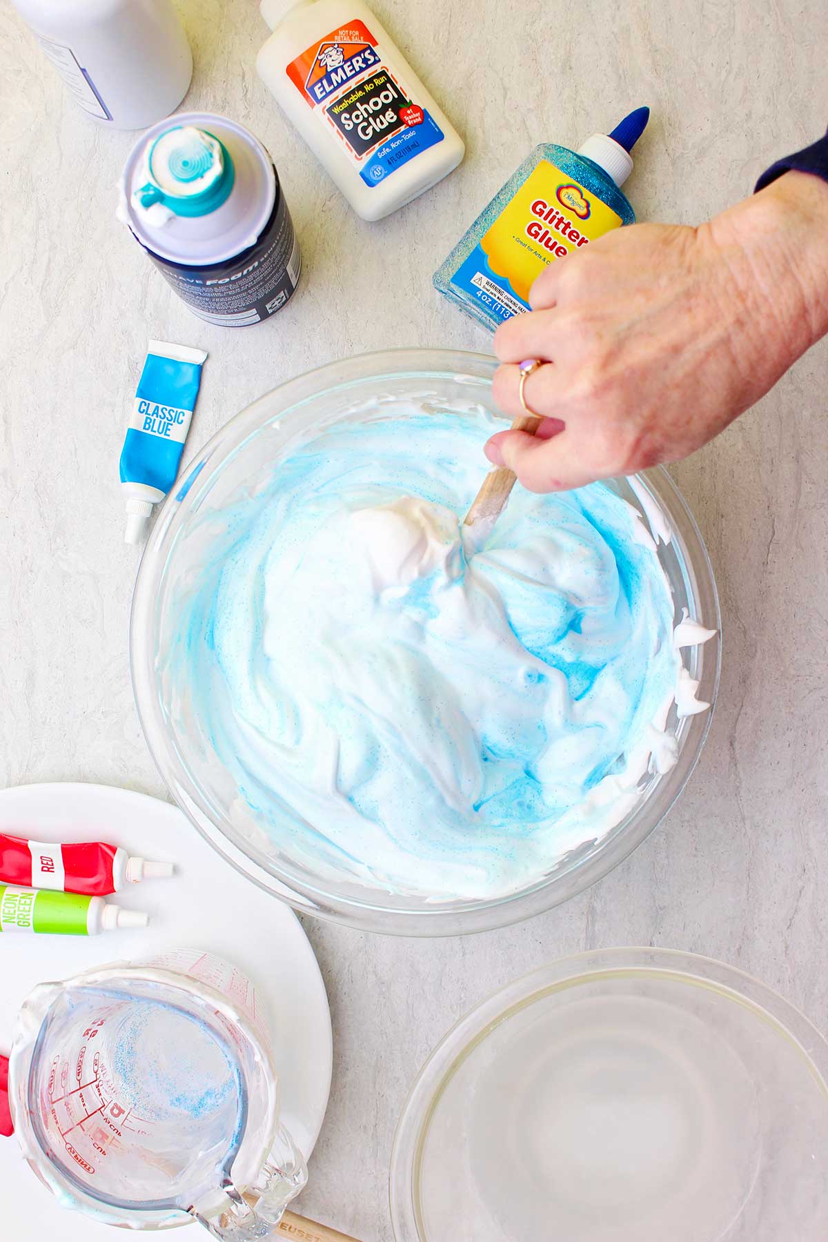 Mão mexendo a corante em um lote de slime fofo azul claro em uma tigela de vidro com suprimentos espalhados ao redor.