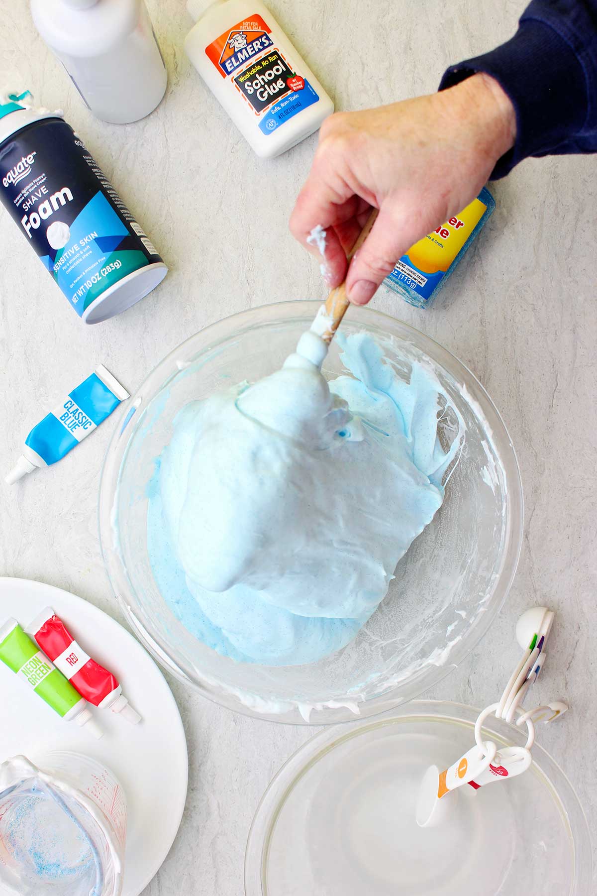 Mão mexendo um lote de slime fofo azul claro em uma tigela de vidro com suprimentos espalhados ao redor.