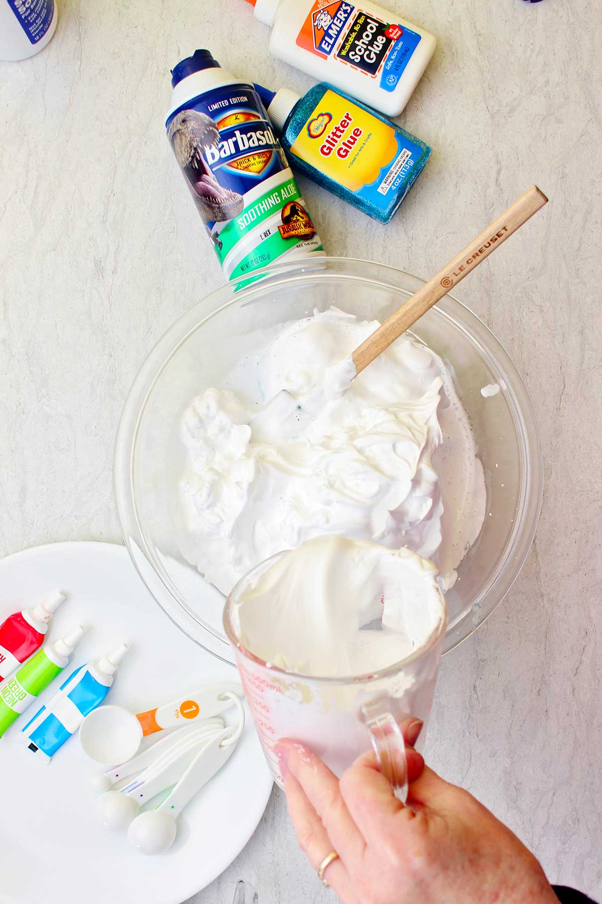 Mão despejando um ingrediente em uma tigela de creme de barbear em uma tigela de vidro com suprimentos para slime fluffy espalhados ao redor.