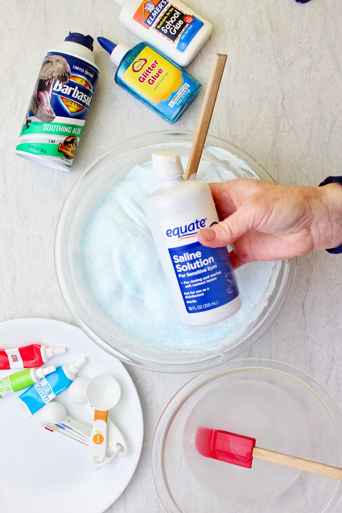 Mão segurando uma garrafa de solução salina acima de uma leva de slime fluffy em uma tigela de vidro com suprimentos espalhados ao redor.