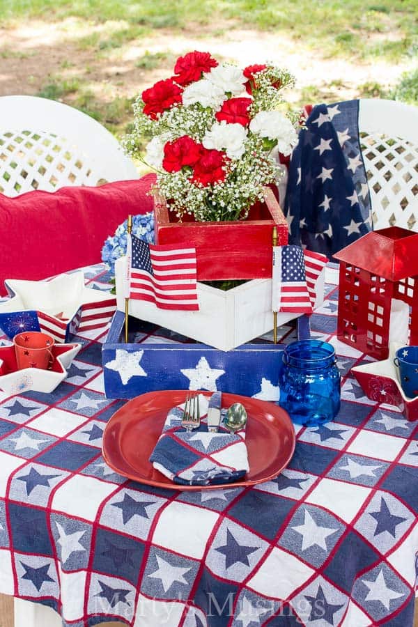 Decorações baratas para a mesa do 4 de Julho, feitas com achados de vendas de garagem, brechós e itens que já temos em casa, com flores da estação, criam um cenário rústico e perfeito ao ar livre. Estrelas vermelhas, brancas e azuis sobre a toalha de mesa com pratos vermelhos e flores