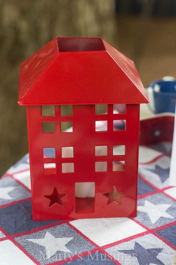 Decorações baratas para a mesa do 4 de Julho, feitas com achados de vendas de garagem, brechós e itens que já temos em casa, com flores da estação, criam um cenário rústico e perfeito ao ar livre. Castiçal decorativo vermelho sobre uma toalha de mesa com estrelas vermelhas, brancas e azuis