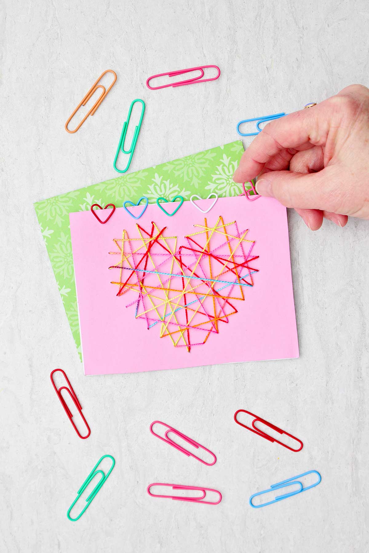 Pessoa colocando um clipe de papel em forma de coração rosa em um cartão de Dia dos Namorados com outros clipes de papel próximos.