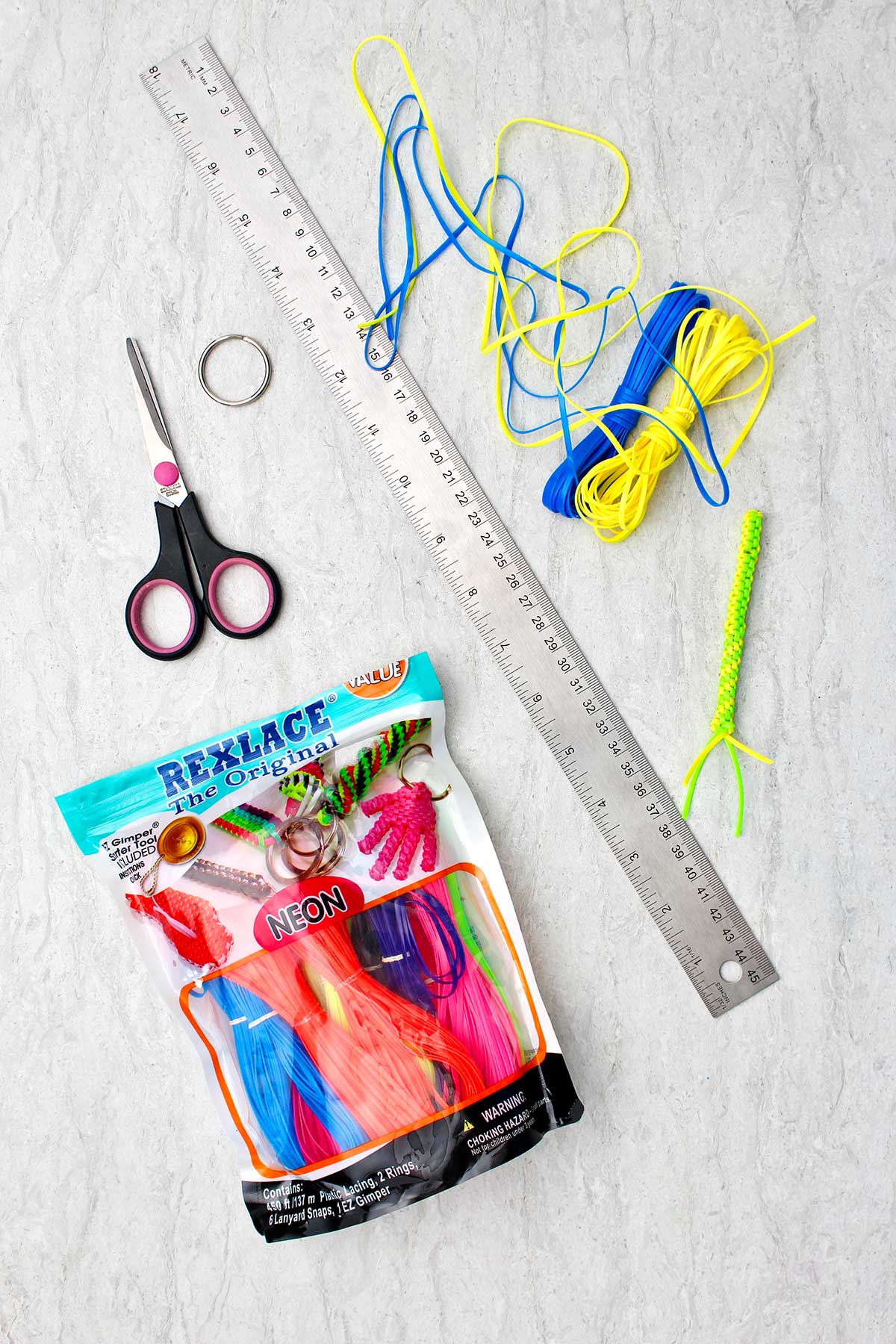 Materiais necessários para fazer um Chaveiro Boondoggle. Rexlace neon, tesoura, uma régua de metal e um chaveiro.