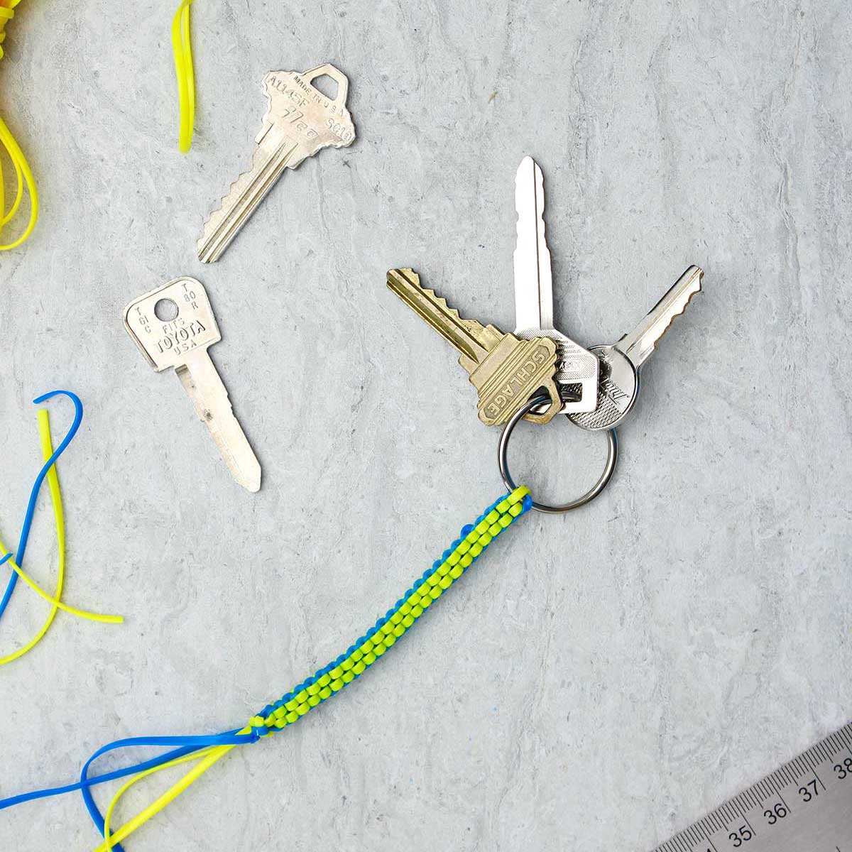 Um chaveiro boondoggle concluído em azul e amarelo com três chaves penduradas e suprimentos próximos.