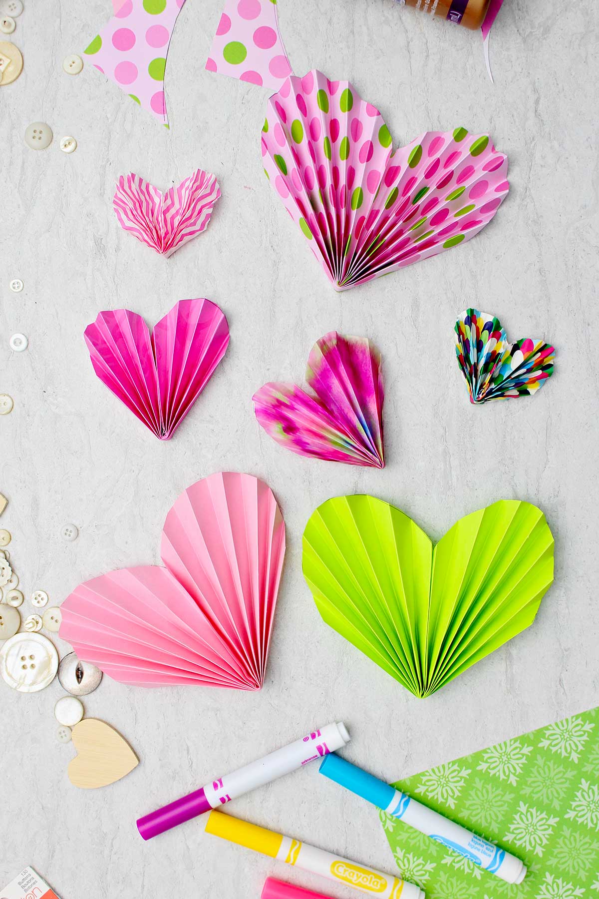 Acordeões de papel rosa e verde usados para decorar cartões e valentins, com algumas canetas e botões espalhados nas proximidades.
