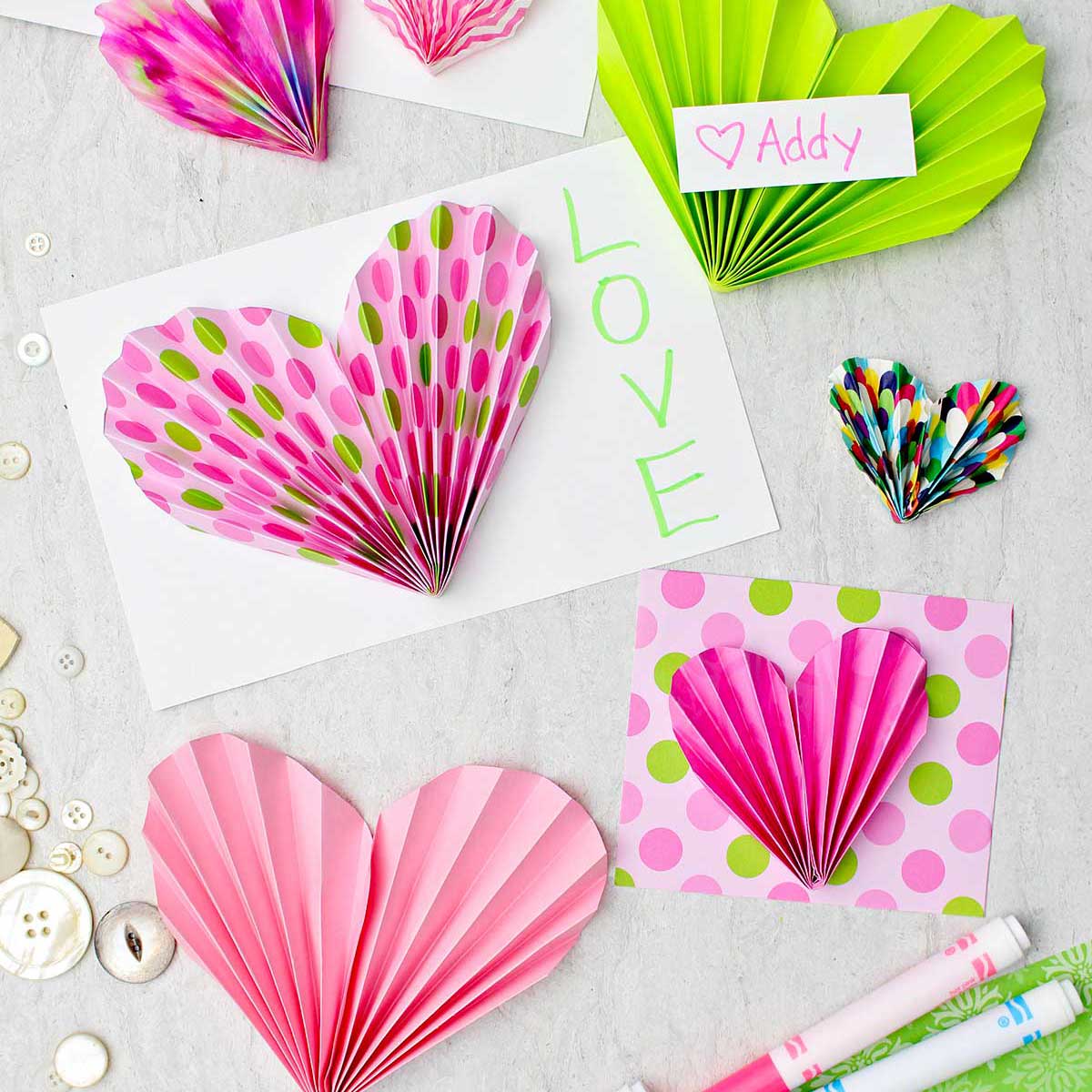 Acordeões de papel rosa e verde usados para decorar cartões e valentins com algumas canetas e botões espalhados nas proximidades.