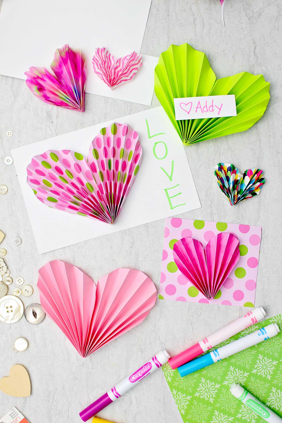 Corações de papel em tons de rosa e verde usados para decorar cartões e valentines com alguns marcadores e botões espalhados nas proximidades.