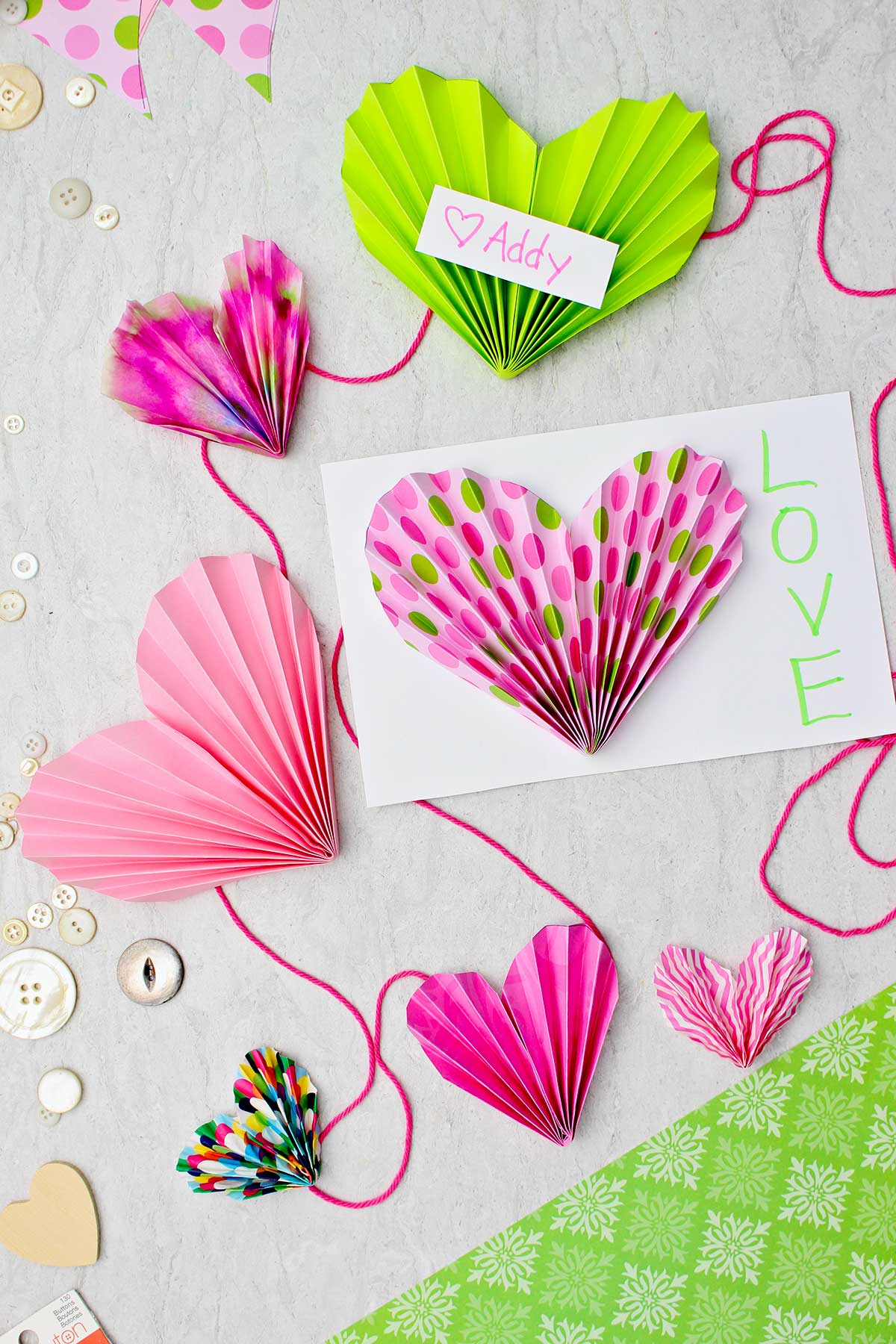 Corações de papel em acordeão rosa e verde completos. Alguns feitos para cartões do Dia dos Namorados e alguns presos a uma corda para fazer uma guirlanda.