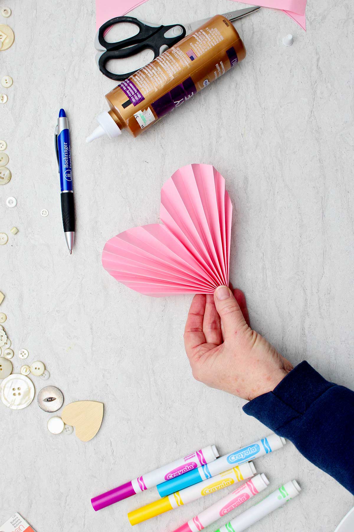 Pessoa segurando um coração de papel em acordeão rosa com outros materiais ao redor.