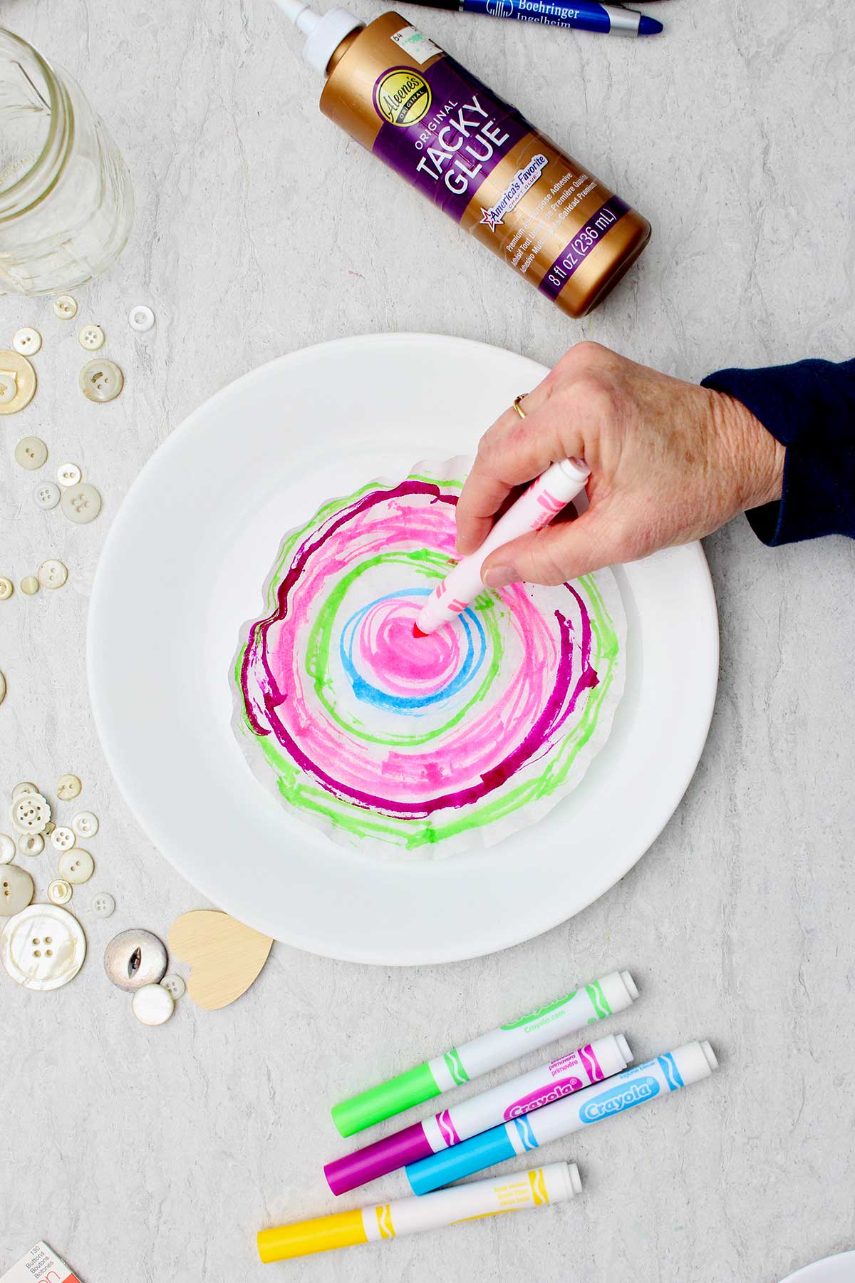 Pessoa colorindo círculos diferentes com caneta para fazer um coração de papel acordeão tie-dye com outros suprimentos por perto.