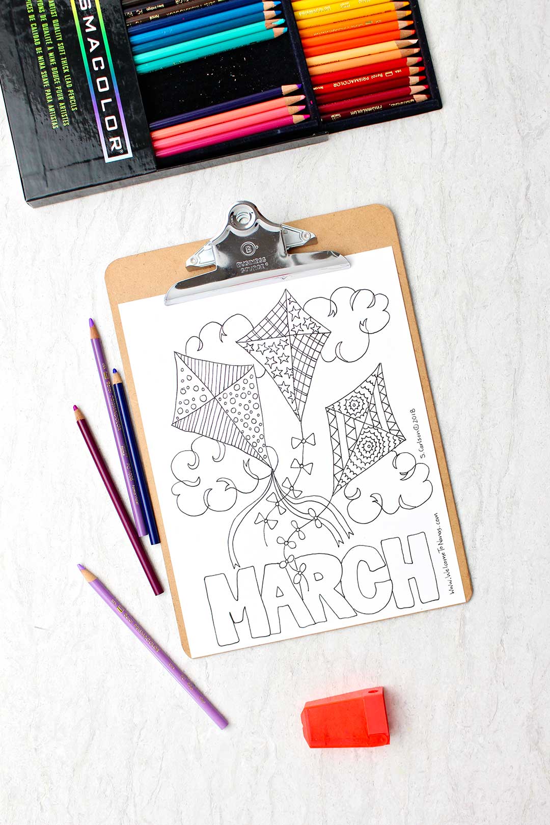 Uma página de colorir de março não colorida em um clipboard com lápis de cor ao lado. A página é uma imagem de três pipas decoradas no céu com a palavra "Março" na parte inferior.