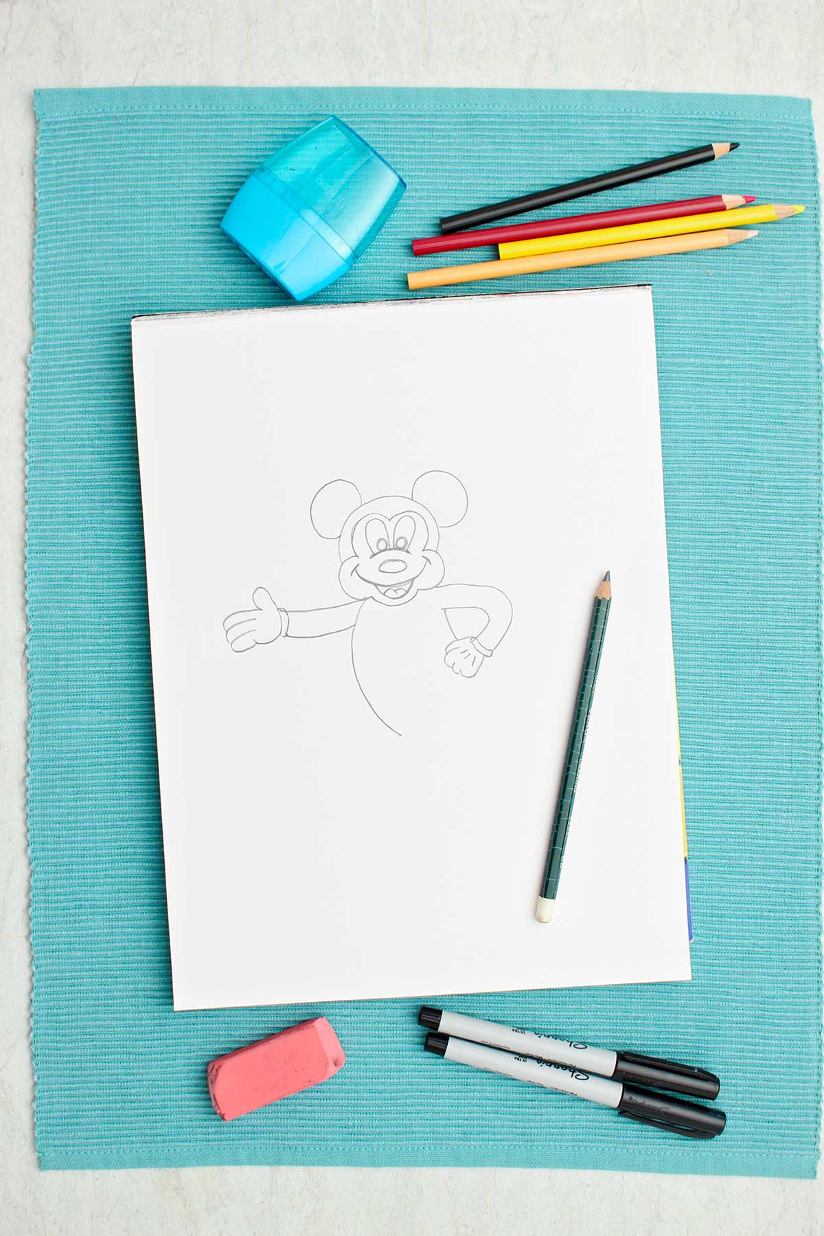 Um esboço a lápis semi-completo do Mickey Mouse e alguns lápis de cor e materiais próximos.