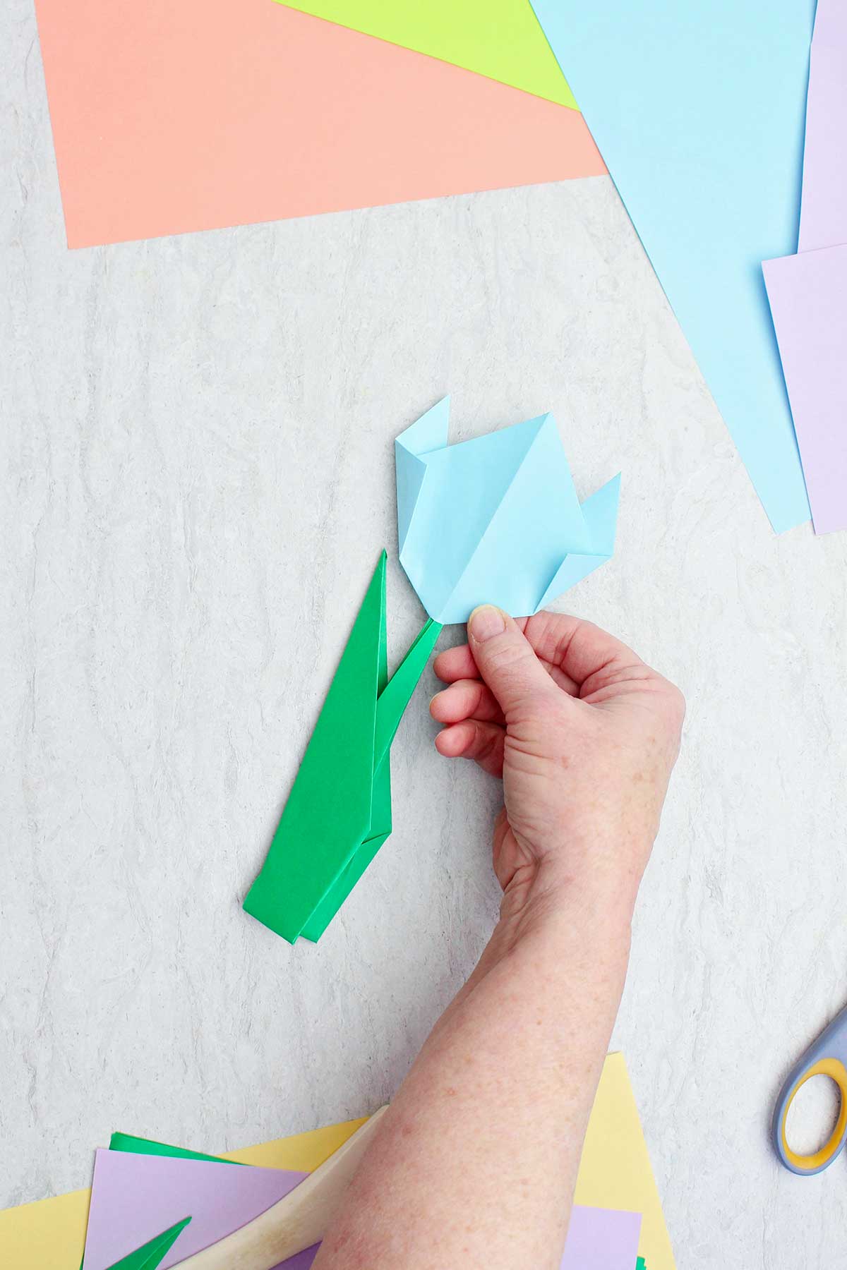 Uma pessoa segurando uma flor de origami azul clara concluída com outros papéis coloridos por perto.