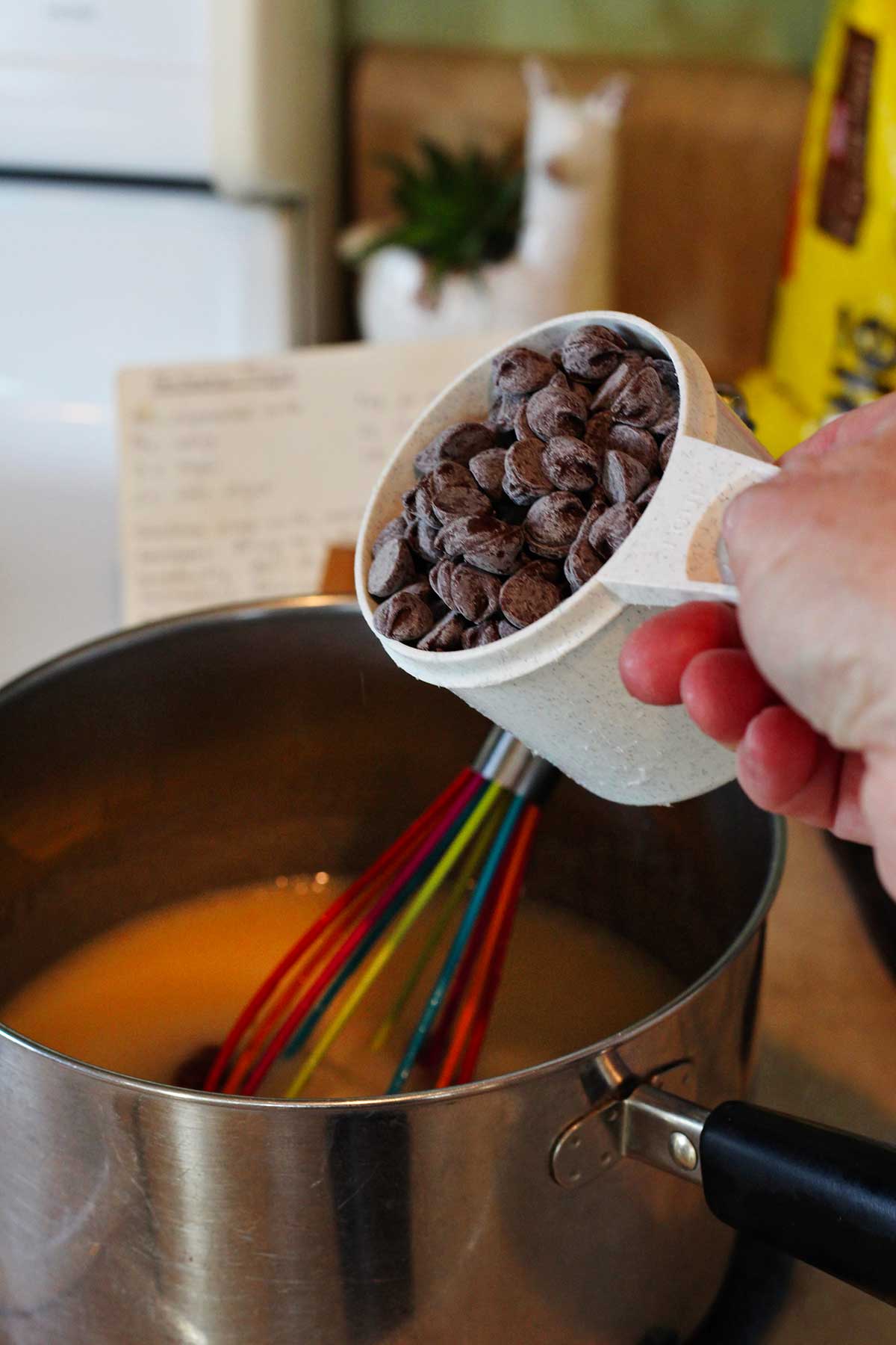 Uma pessoa prestes a adicionar uma xícara de gotas de chocolate à mistura de fudge na panela.