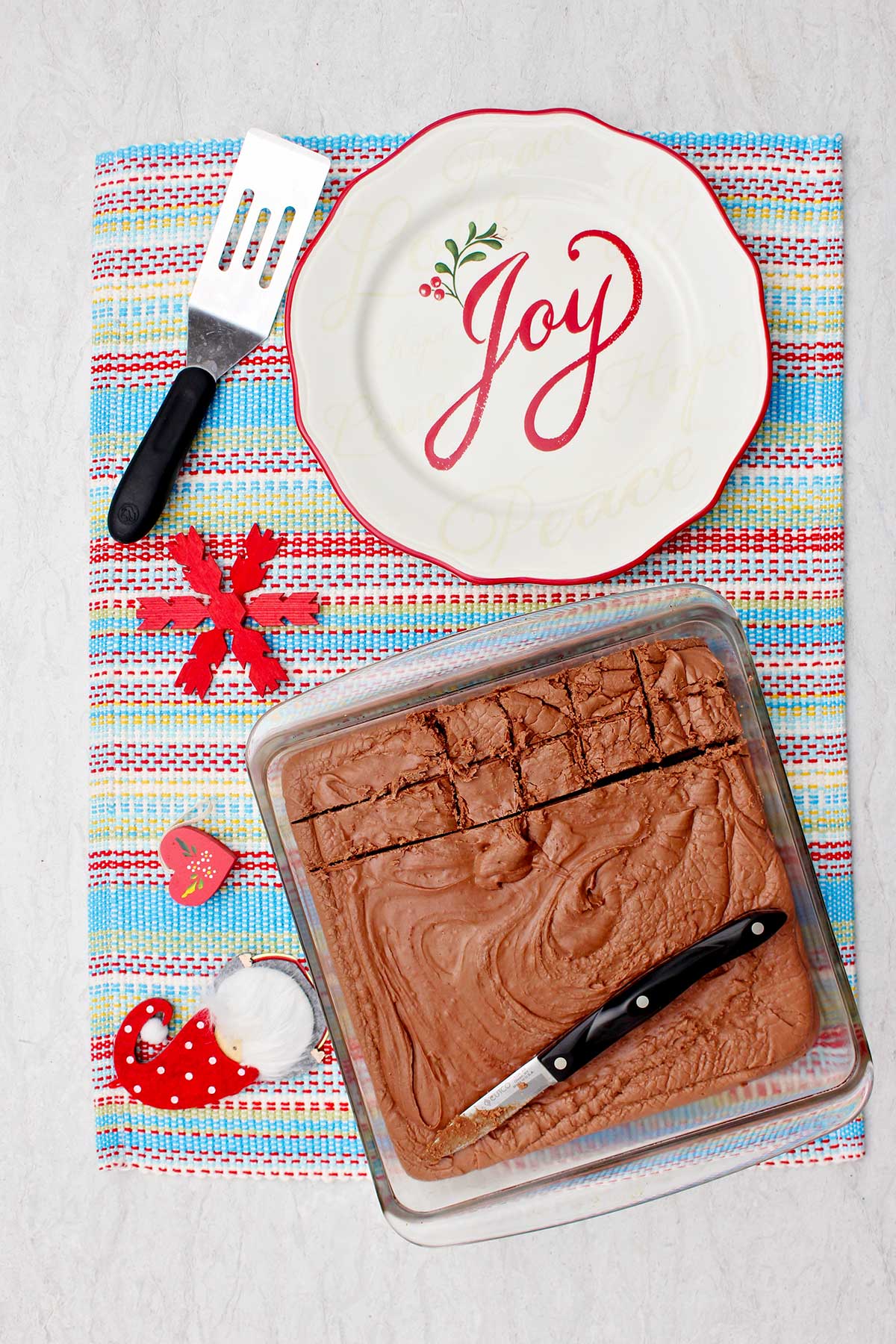 Fudge final em uma forma de vidro sobre um jogo de mesa colorido com um prato festivo e uma espátula ao lado.