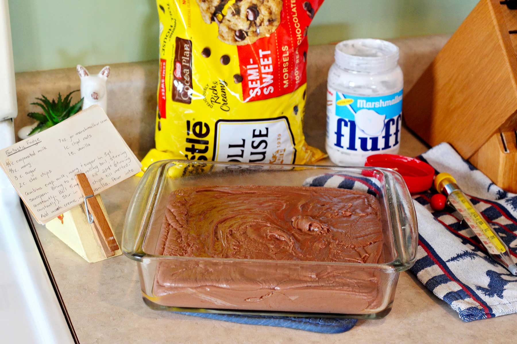 Mistura de fudge na forma de vidro sobre a bancada com chips de chocolate, fluff e receita perto.