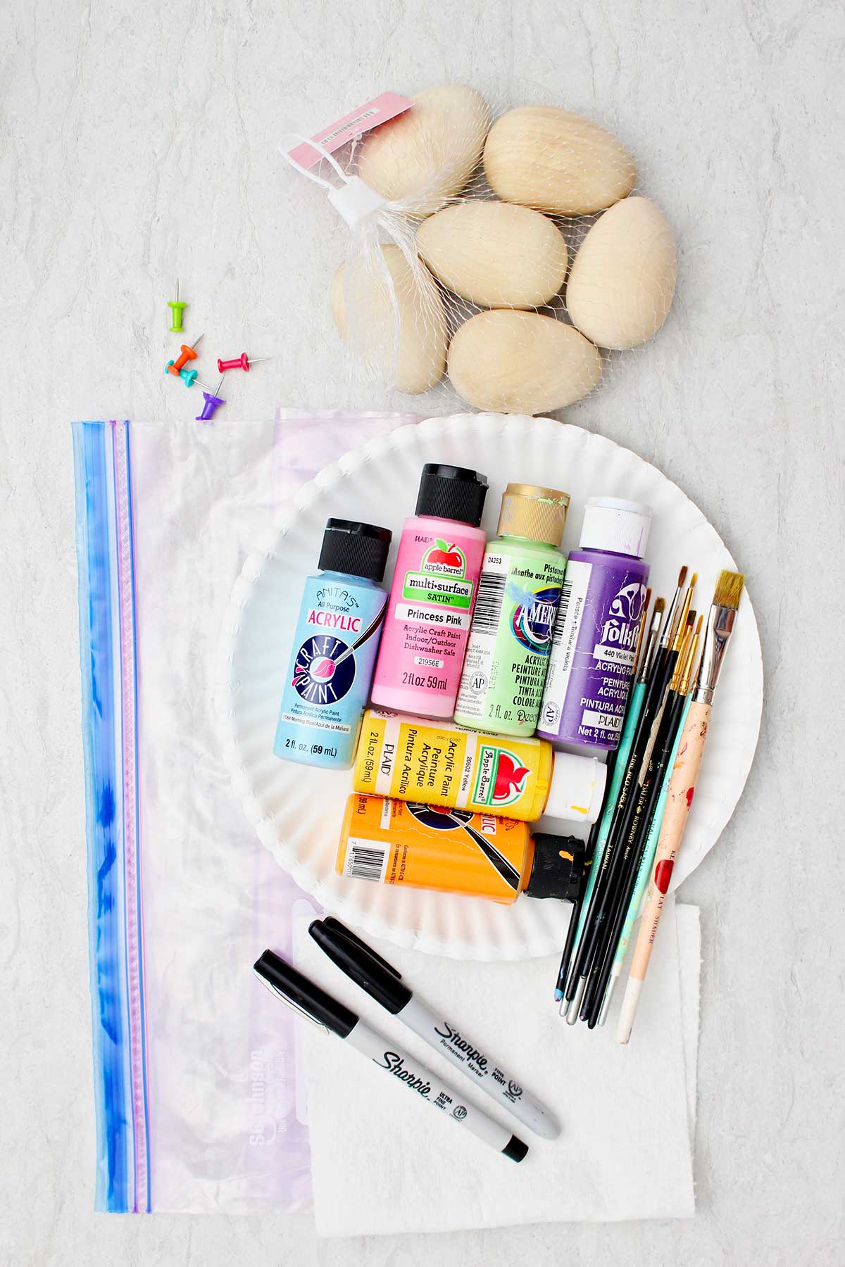Uma bolsa com ovos de madeira, um saco plástico com zip-lock, tintas acrílicas coloridas, pincéis e marcadores sharpie.