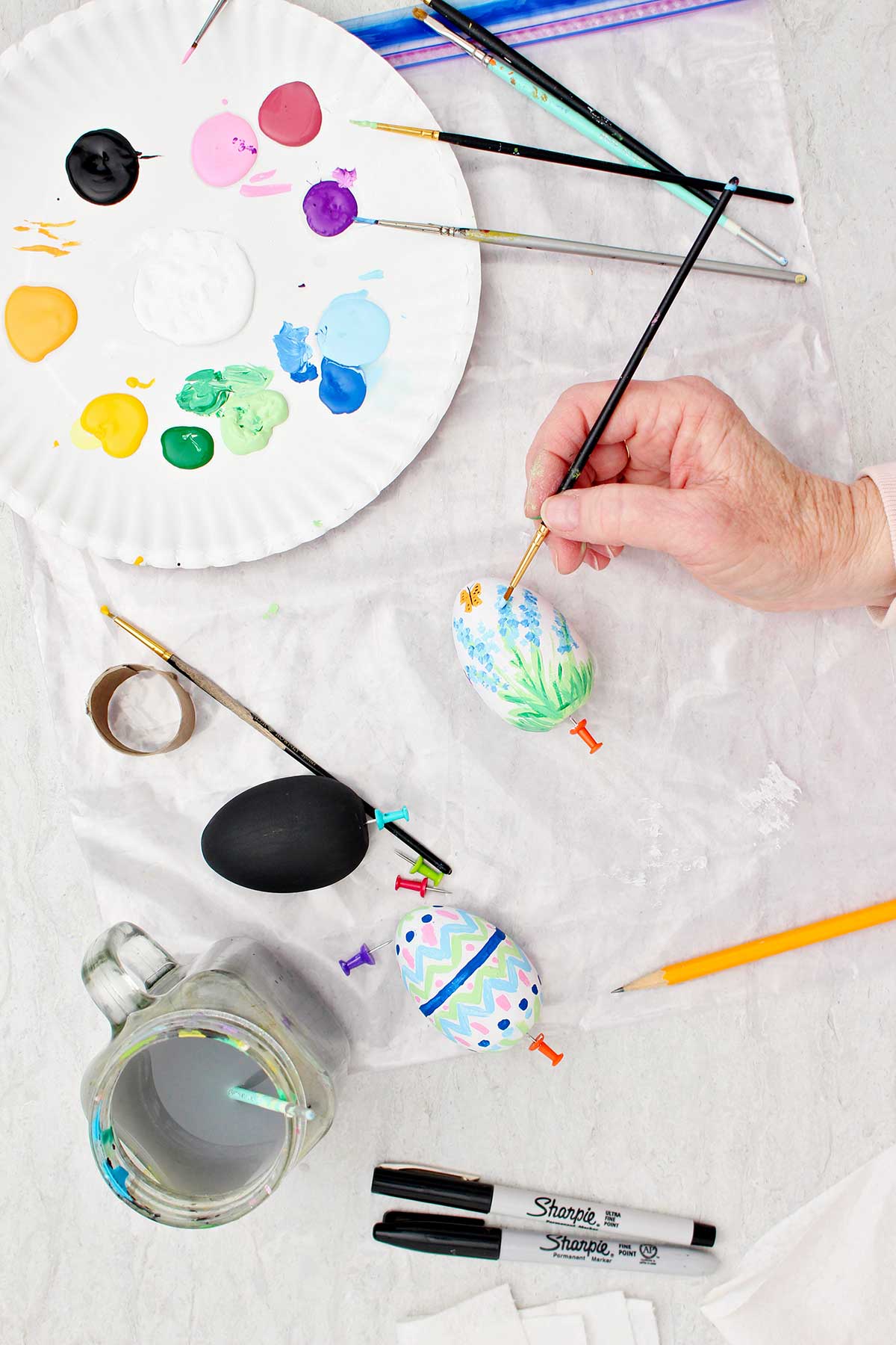 Pessoa colocando detalhes em uma flor azul de um ovo de madeira branco pintado com suprimentos de pintura ao redor.