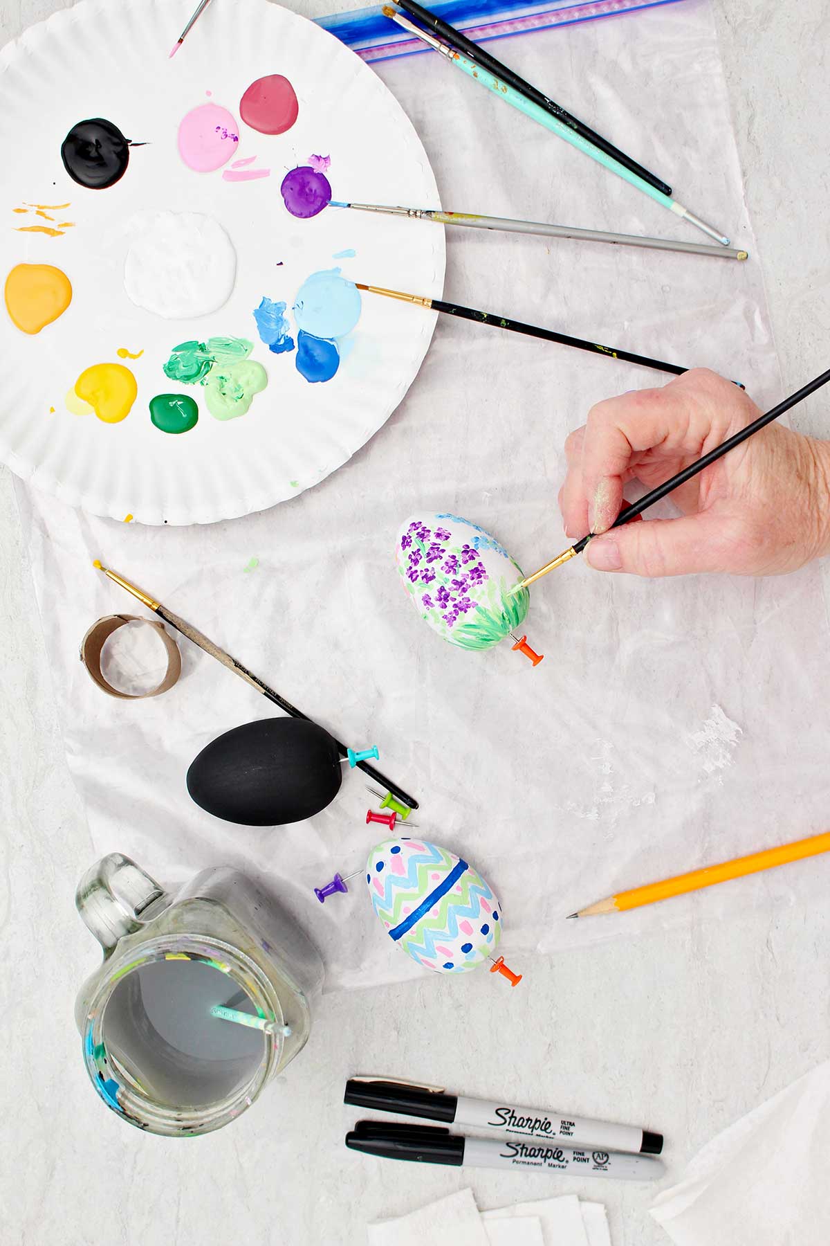 Pessoa colocando detalhes na grama de um ovo de madeira branco pintado com flores, com suprimentos de pintura ao redor.