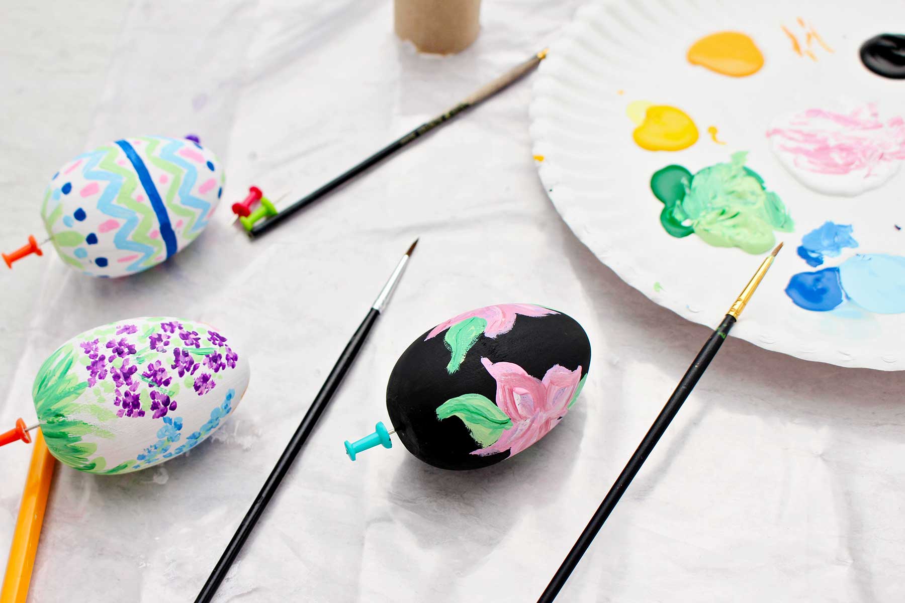 Ovos de madeira pintados com flores pretas, flores brancas e padrões secando com materiais de pintura por perto.