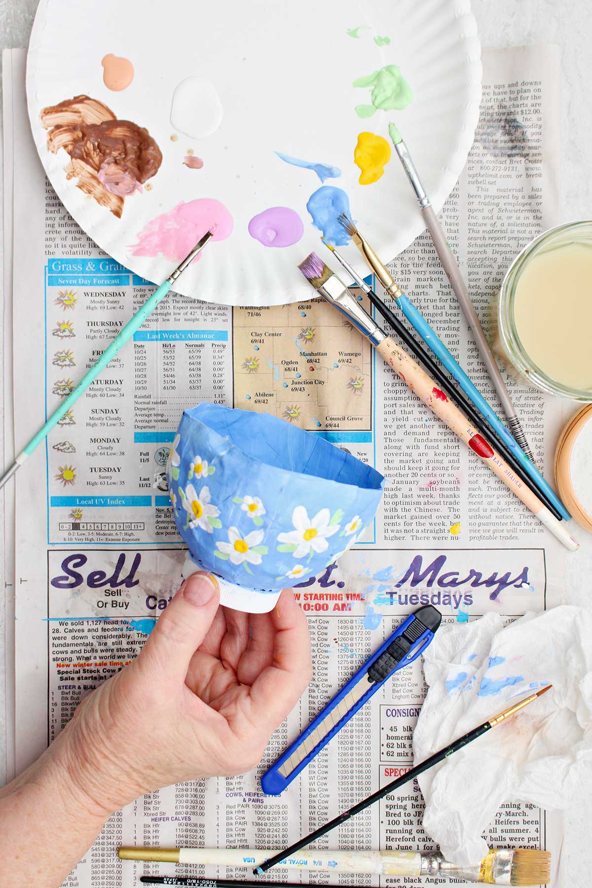 Uma pessoa segurando um projeto de papel machê pintado de fundo azul claro com flores brancas e amarelas.