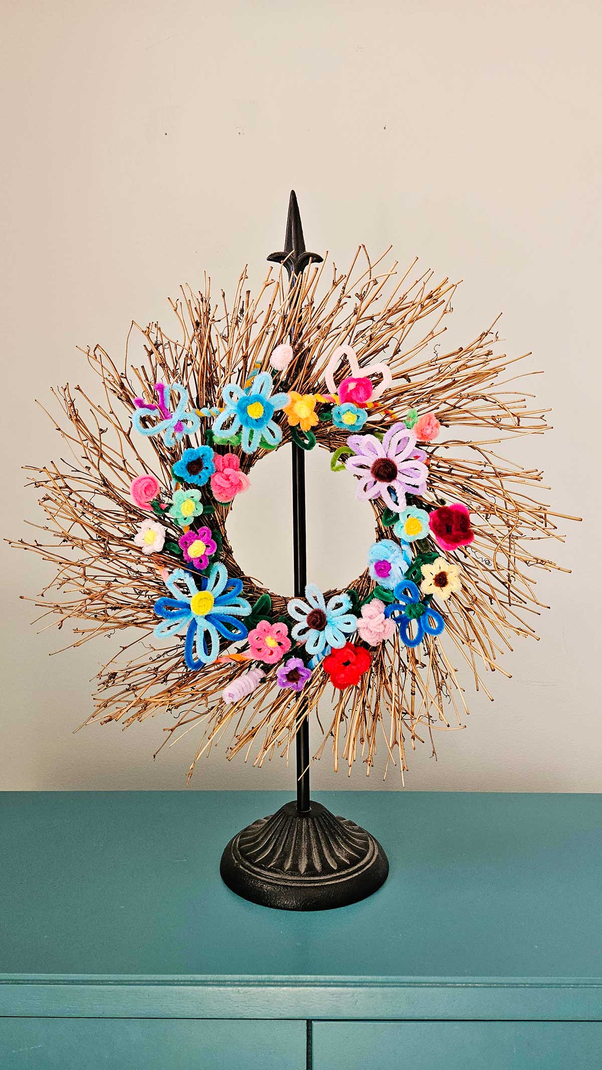 Uma guirlanda de galhos em um suporte com flores de limpador de pipe multicoloridas presas a ela.