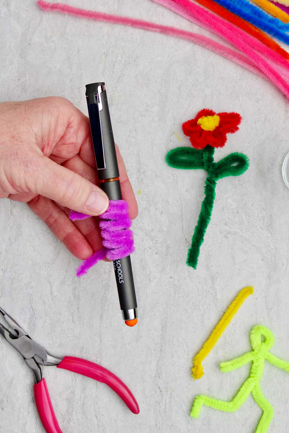 Uma pessoa segurando uma caneta com um limpador de pipe roxo enrolado ao seu redor, com uma flor de limpador de pipe e outros limpadores de pipe ao fundo.
