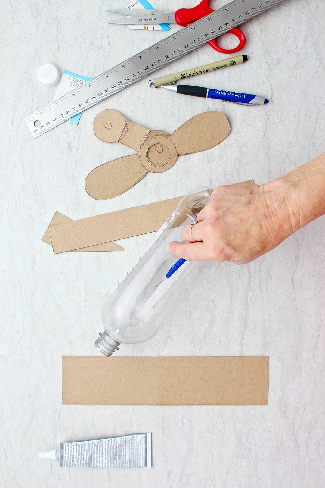Mão usando um cortador para fazer um corte na garrafa plástica vazia com asas de papelão e hélices próximas.